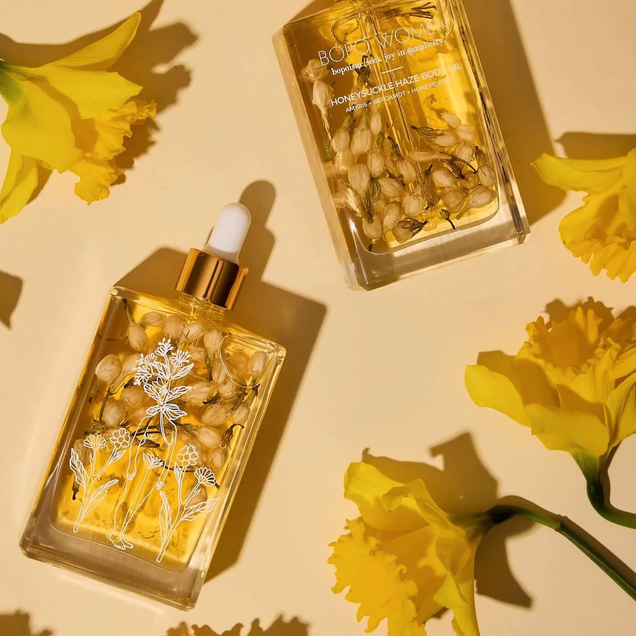 Bopo Women Body Oil - Honeysuckle Haze bottles surrounded by yellow flowers, showcasing elegant packaging for a hydrating body oil.