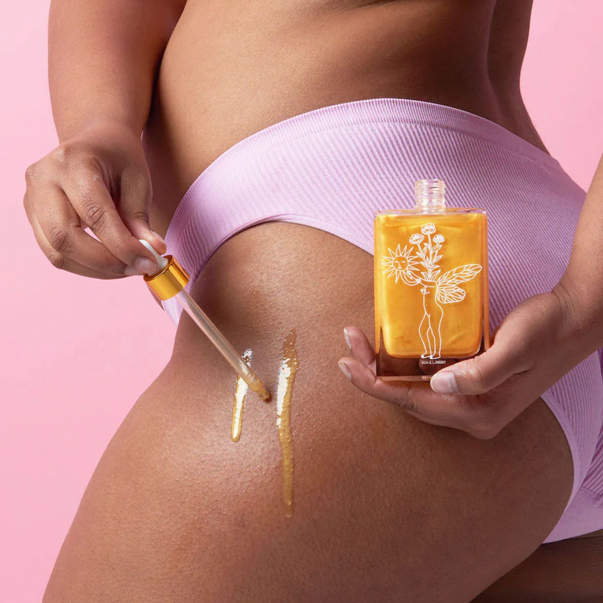 Person applying a golden oil from a dropper bottle to their skin against a pink background