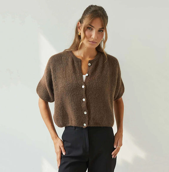 A woman modeling a brown short-sleeve fuzzy knit cardigan with a button-up front, paired with black trousers, against a light off-white wall with soft shadows.