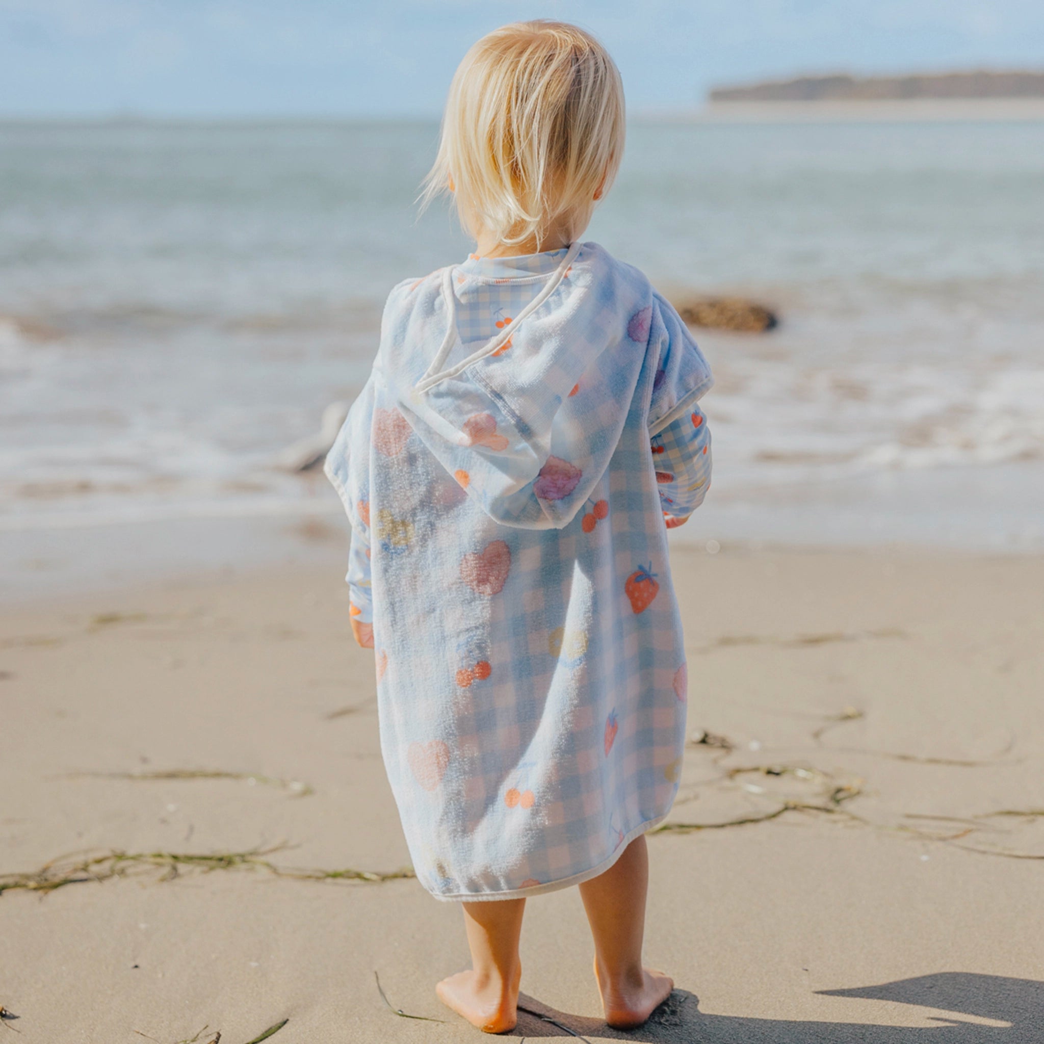 Child in a light blue towel with colorful patterns standing on a sandy beach.