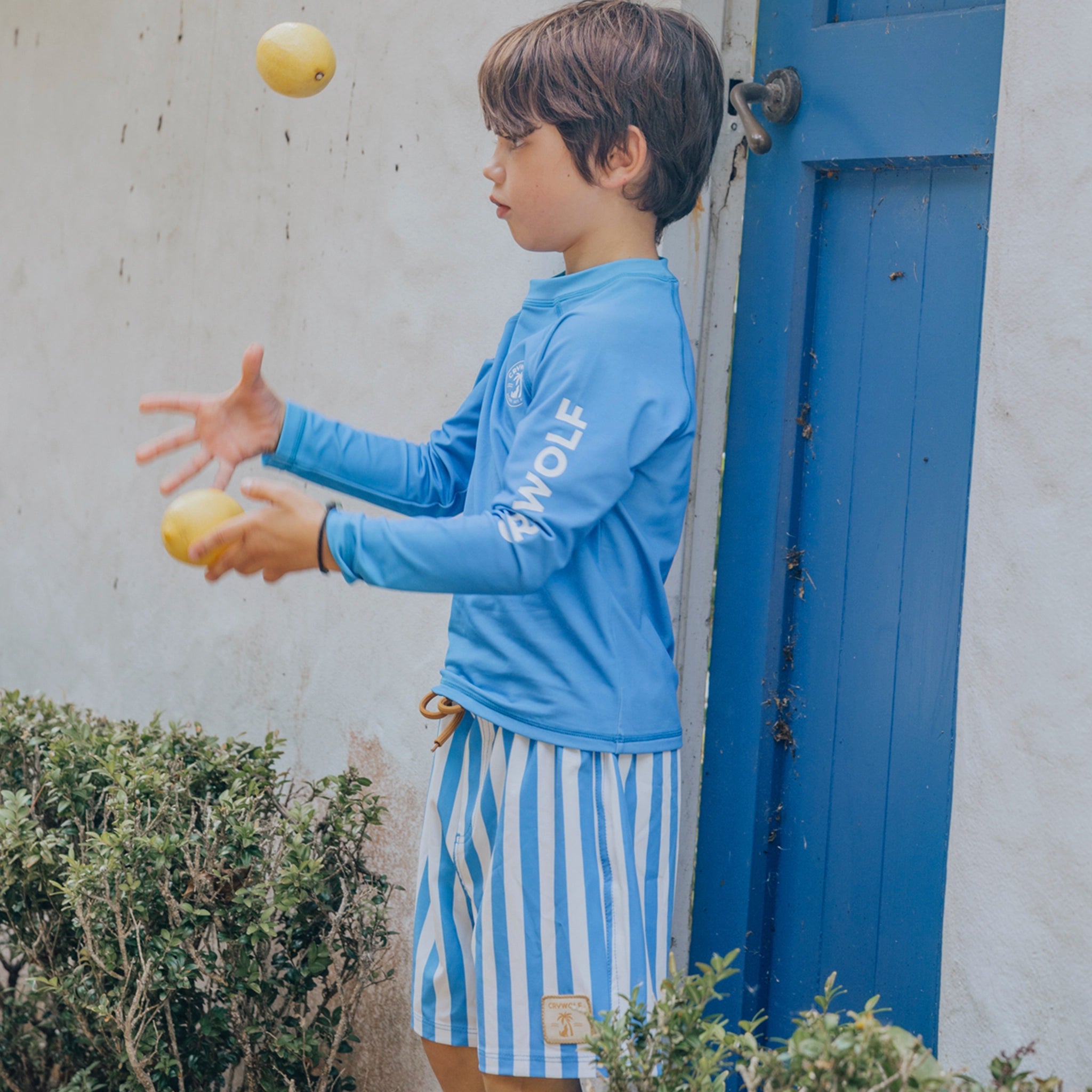 Child in a blue shirt with 'WOLF' logo juggling apples near a blue door.