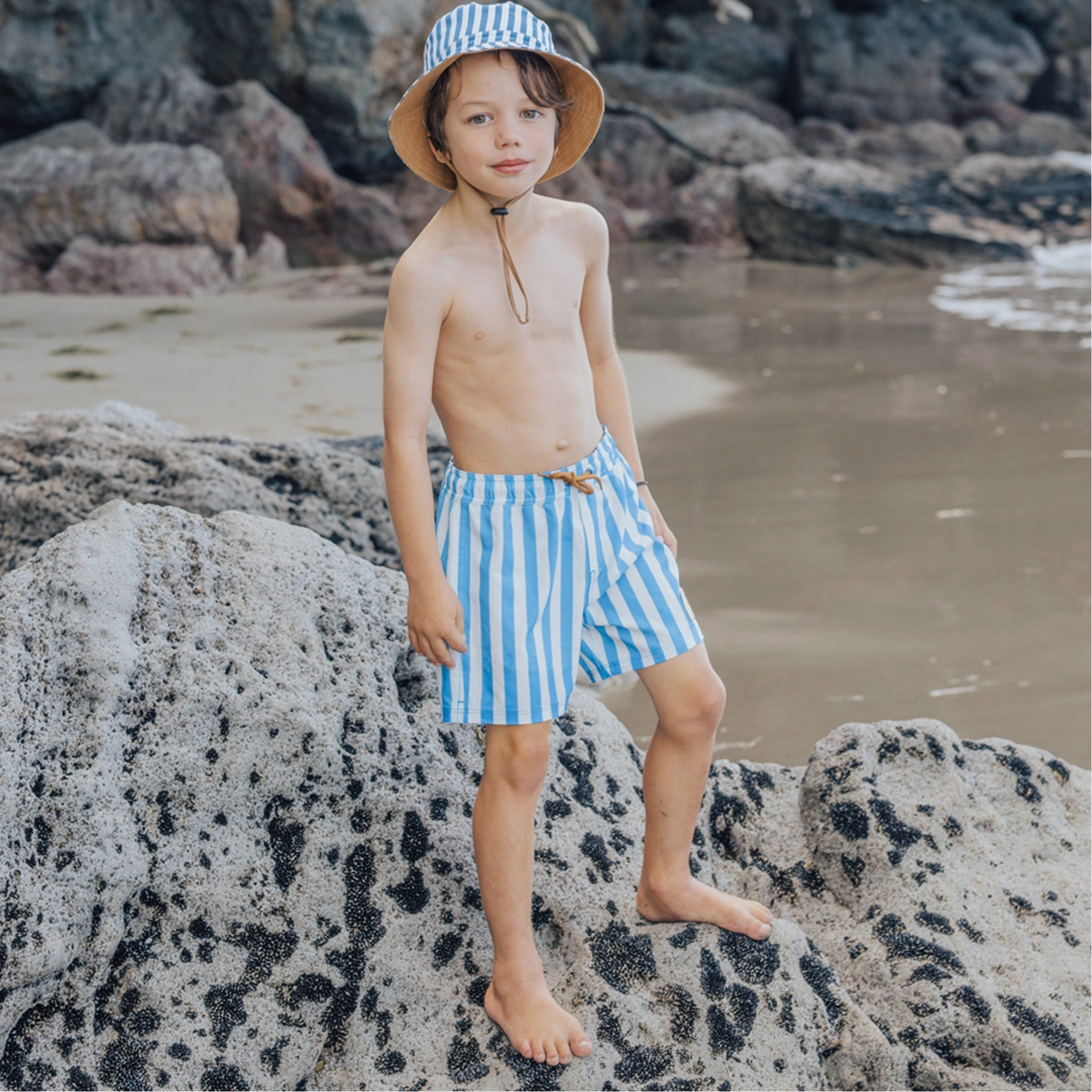 Child wearing blue and white striped swim shorts and a hat on a rocky beach.