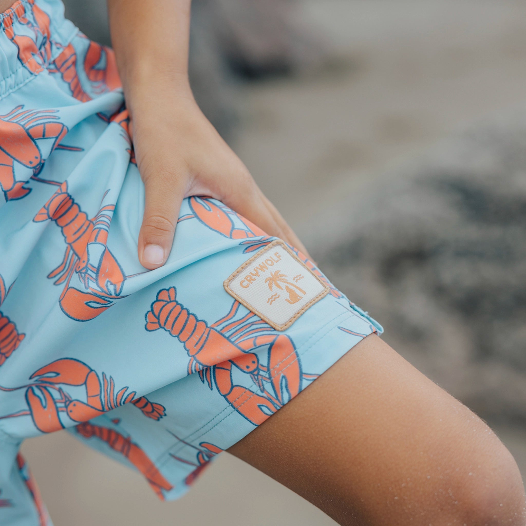 Shorts with lobster pattern held by a person on a blurred beach background