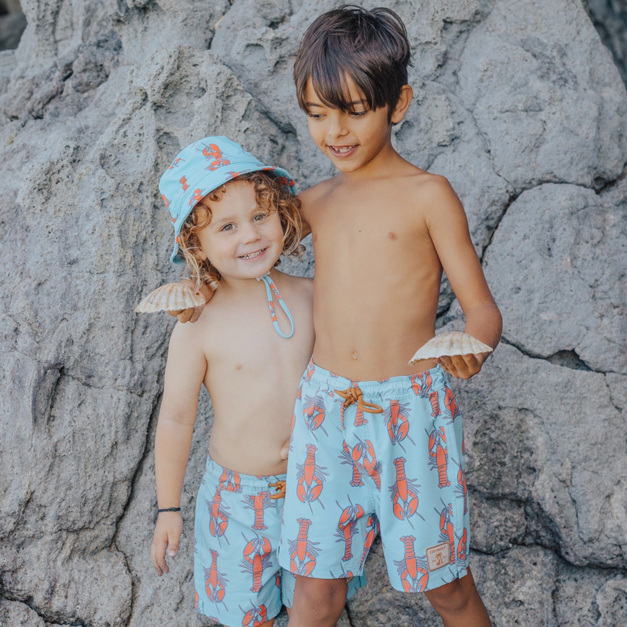 Two children wearing matching blue swim trunks with orange patterns against a rocky background.