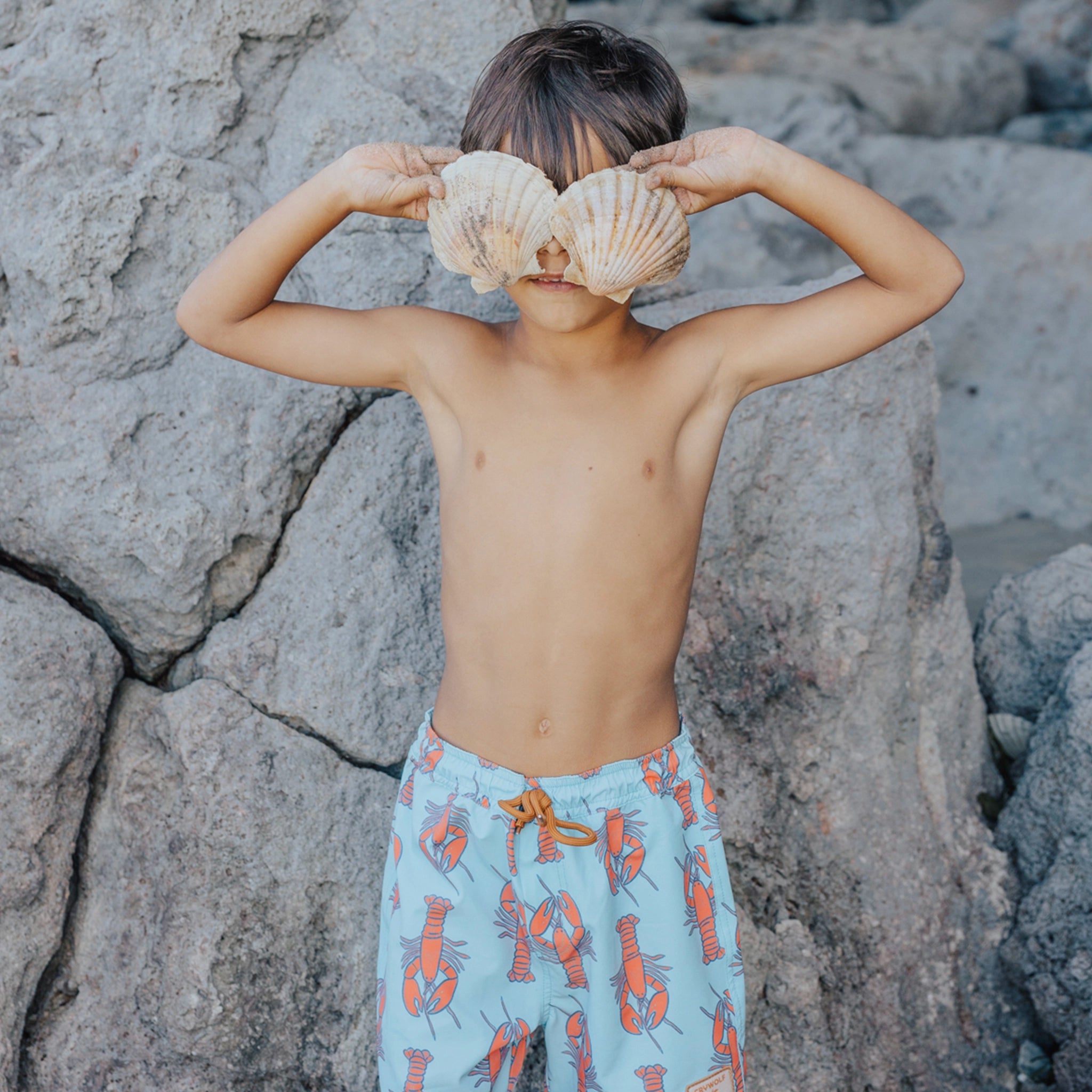 Child holding seashells over their eyes against a rocky background