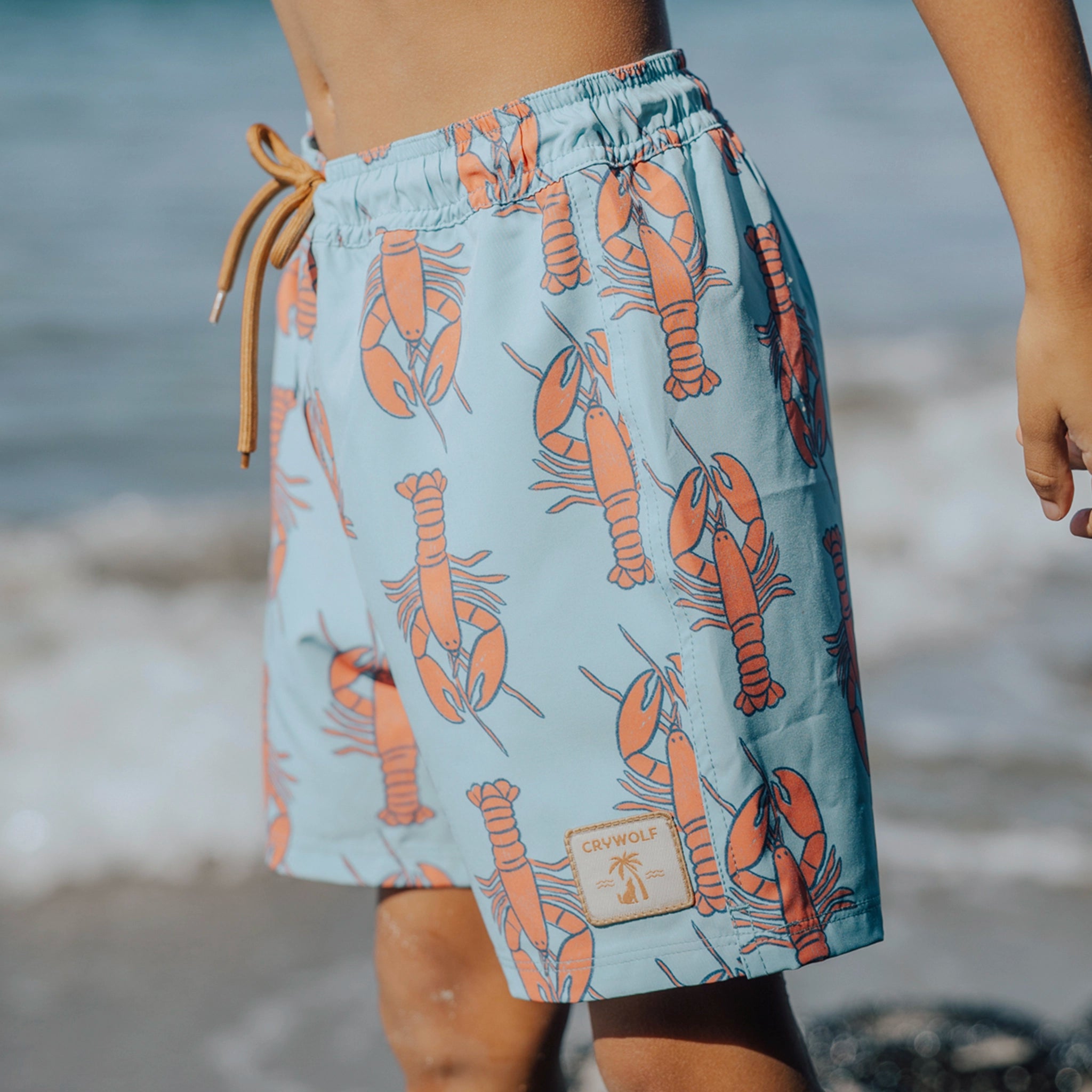 Person wearing a blue swim shorts with orange lobster pattern on a beach.