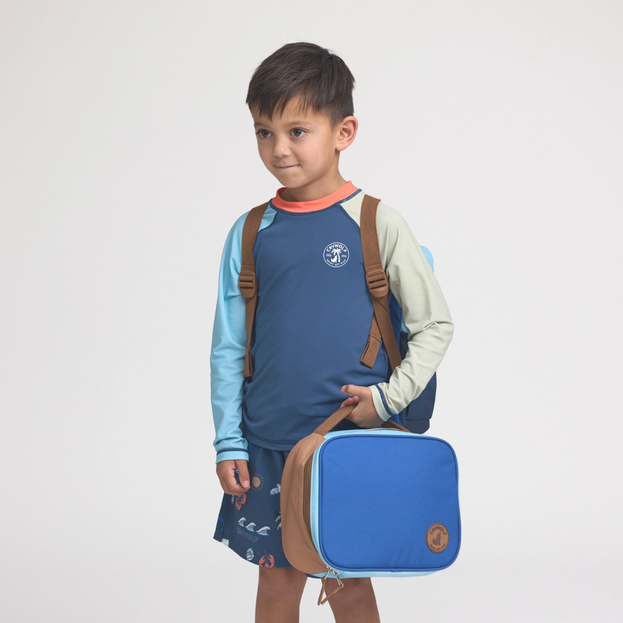 Child wearing a blue shirt with a backpack holding a blue lunch bag on a white background
