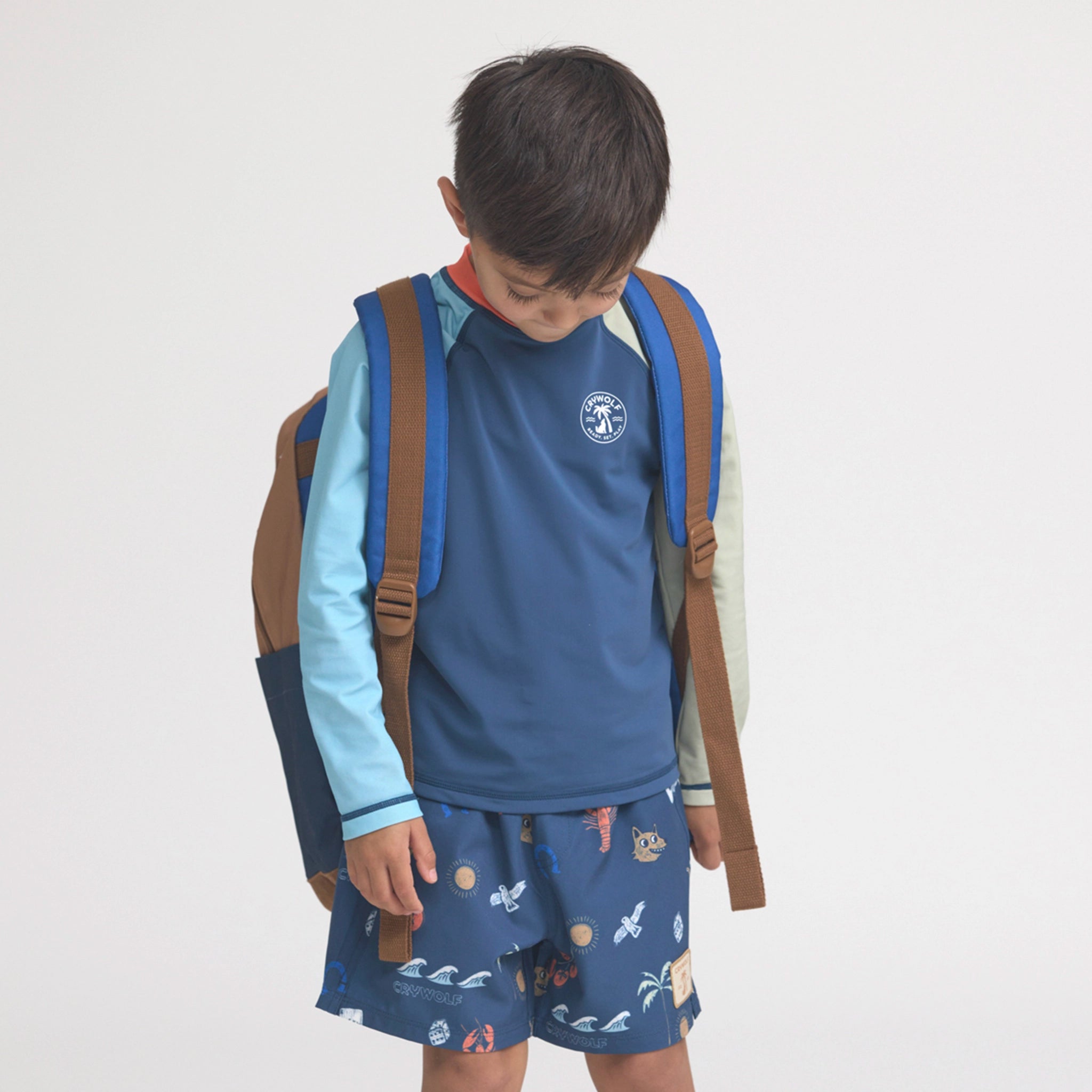 Child wearing a blue shirt with a logo, patterned shorts, and a brown backpack on a light gray background