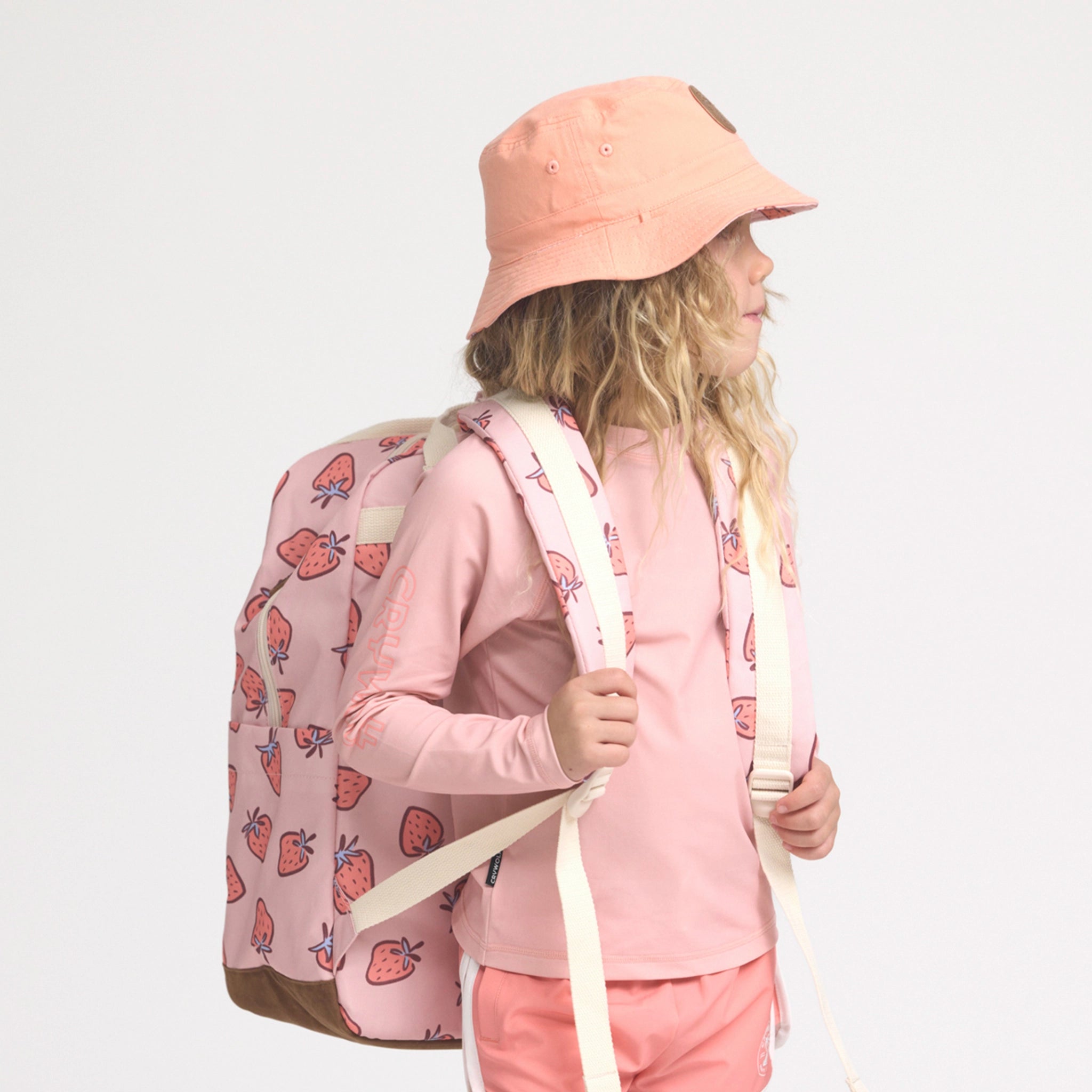 Child wearing a pink outfit with a strawberry-patterned backpack on a light gray background