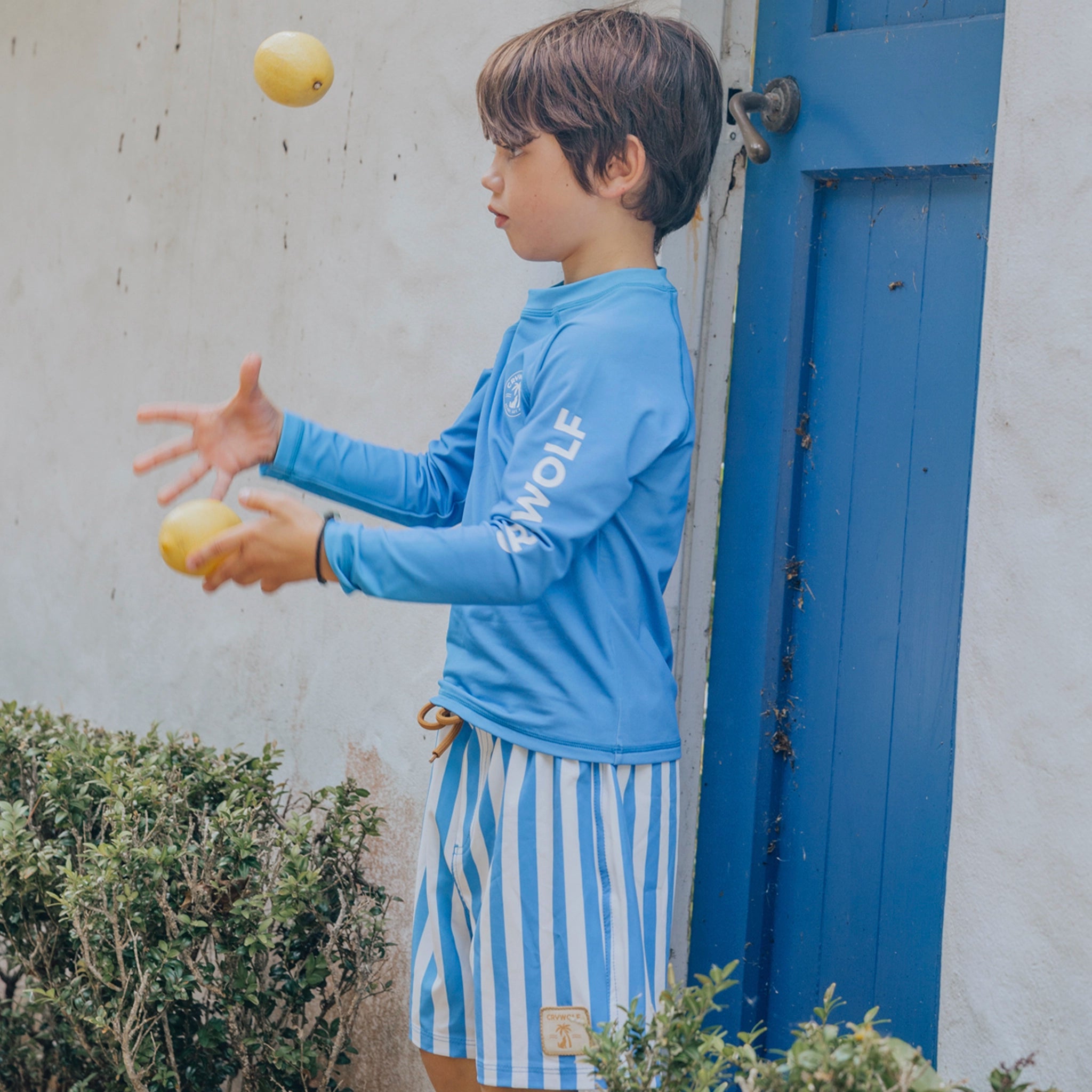 Child in blue rash vest and striped shorts juggling balls outdoors.