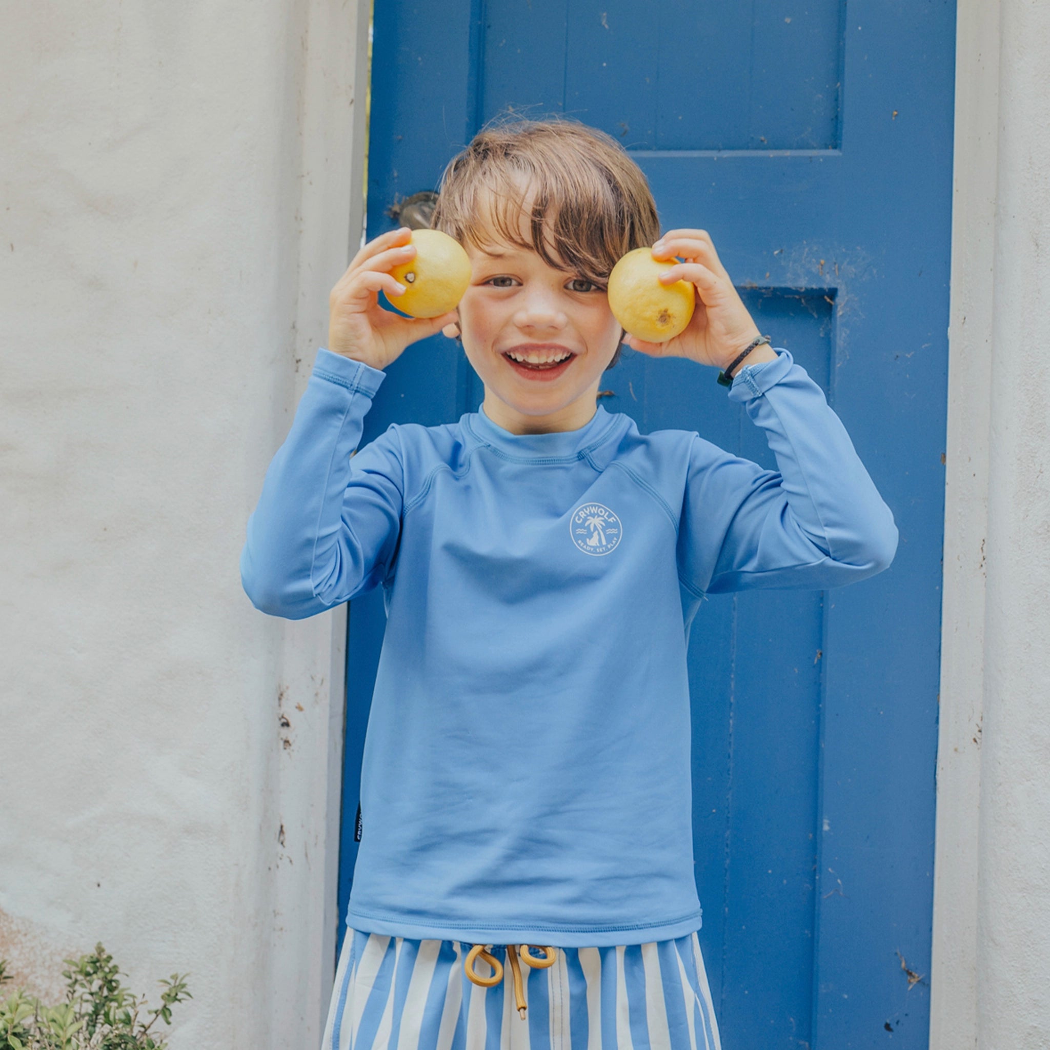 Child wearing a blue long-sleeve rash vest with a logo, holding lemons in front of a blue door.