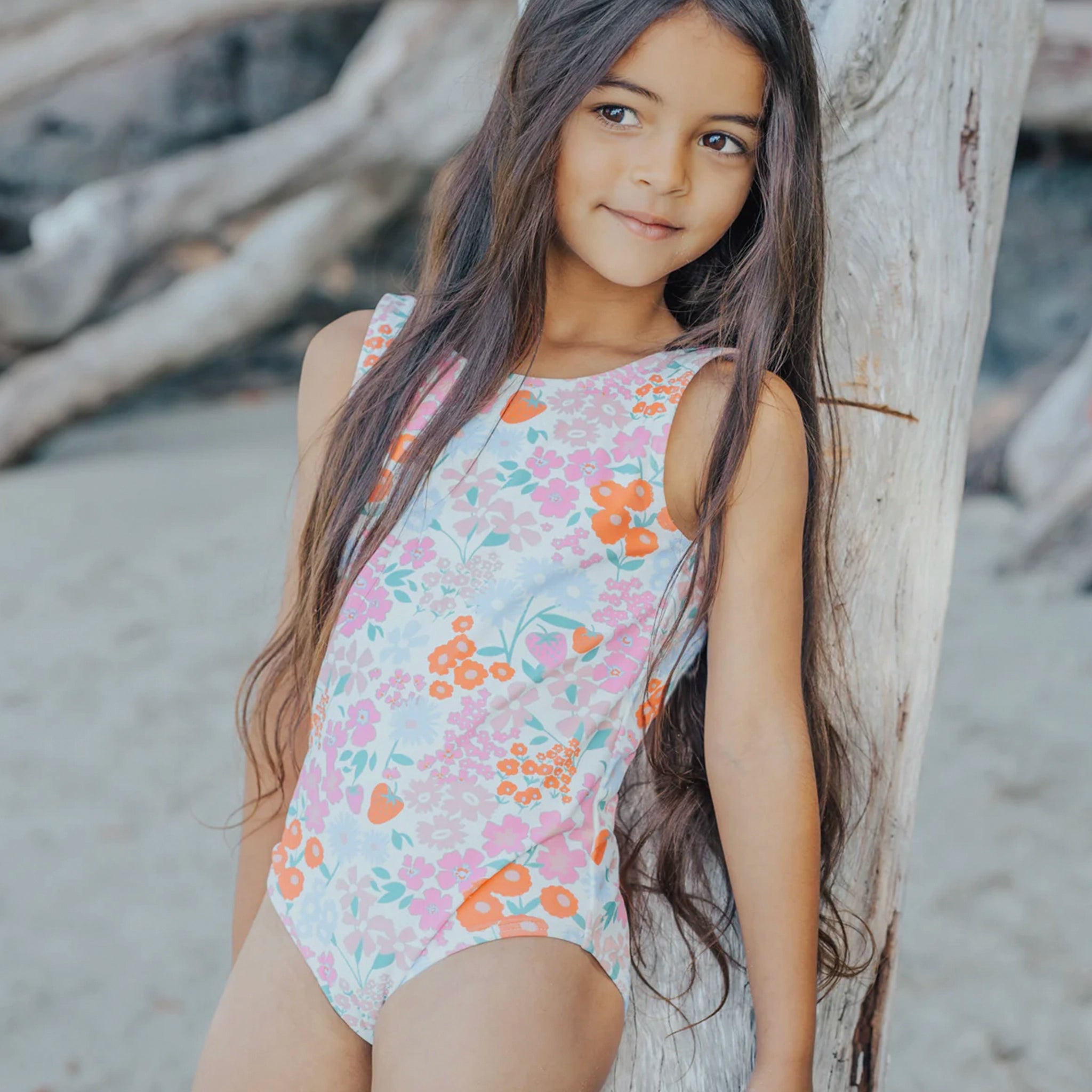 Young girl in a floral swimsuit standing next to a tree on a beach.