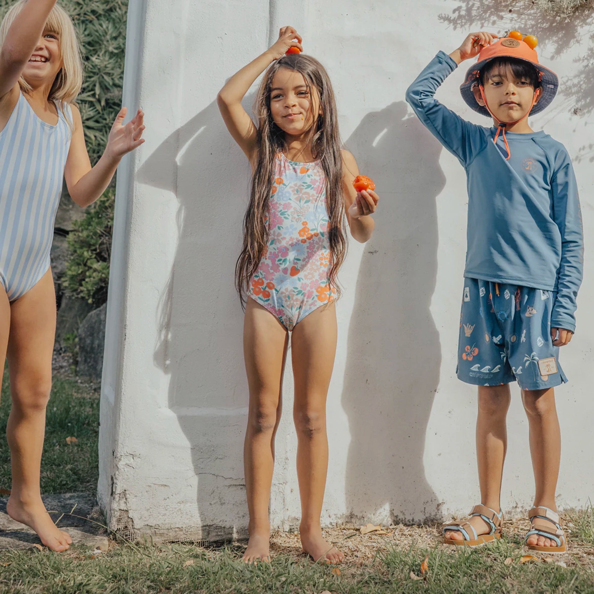 Three children in swimsuits standing against a white wall outdoors.