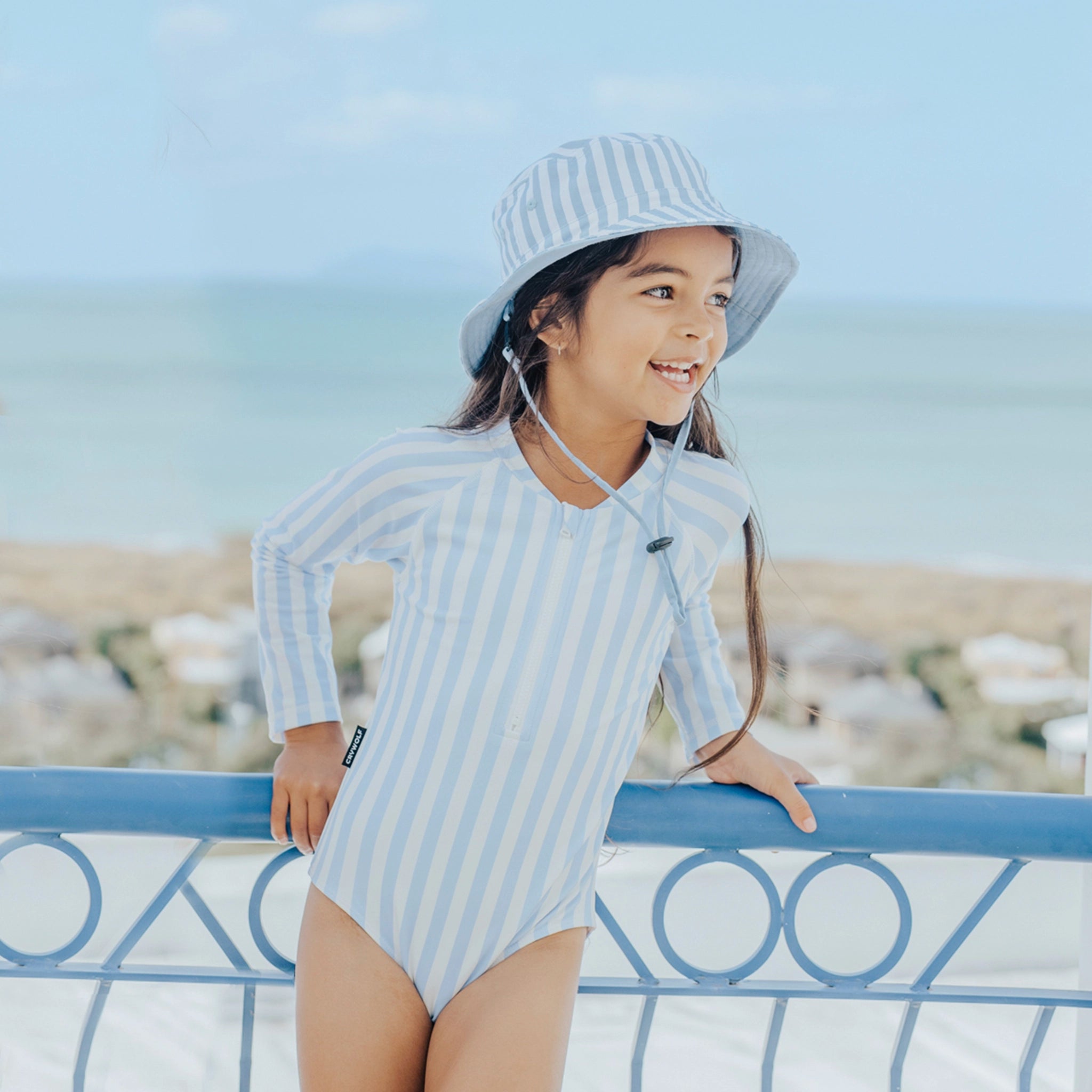 Child wearing a blue and white striped swimsuit and hat, standing on a balcony with a scenic background.