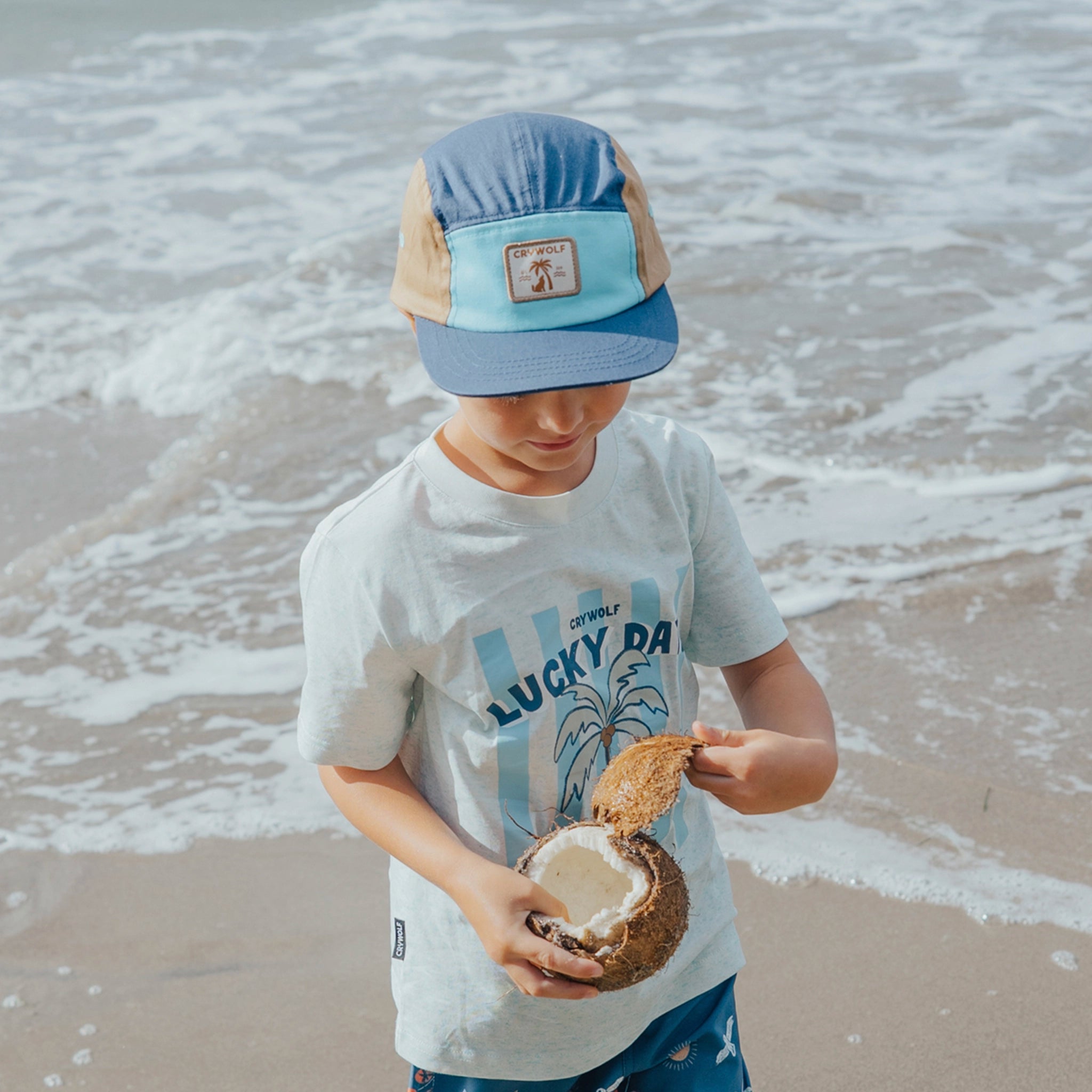 Child on a beach holding a coconut, wearing a blue cap and 'Lucky Day' t-shirt.