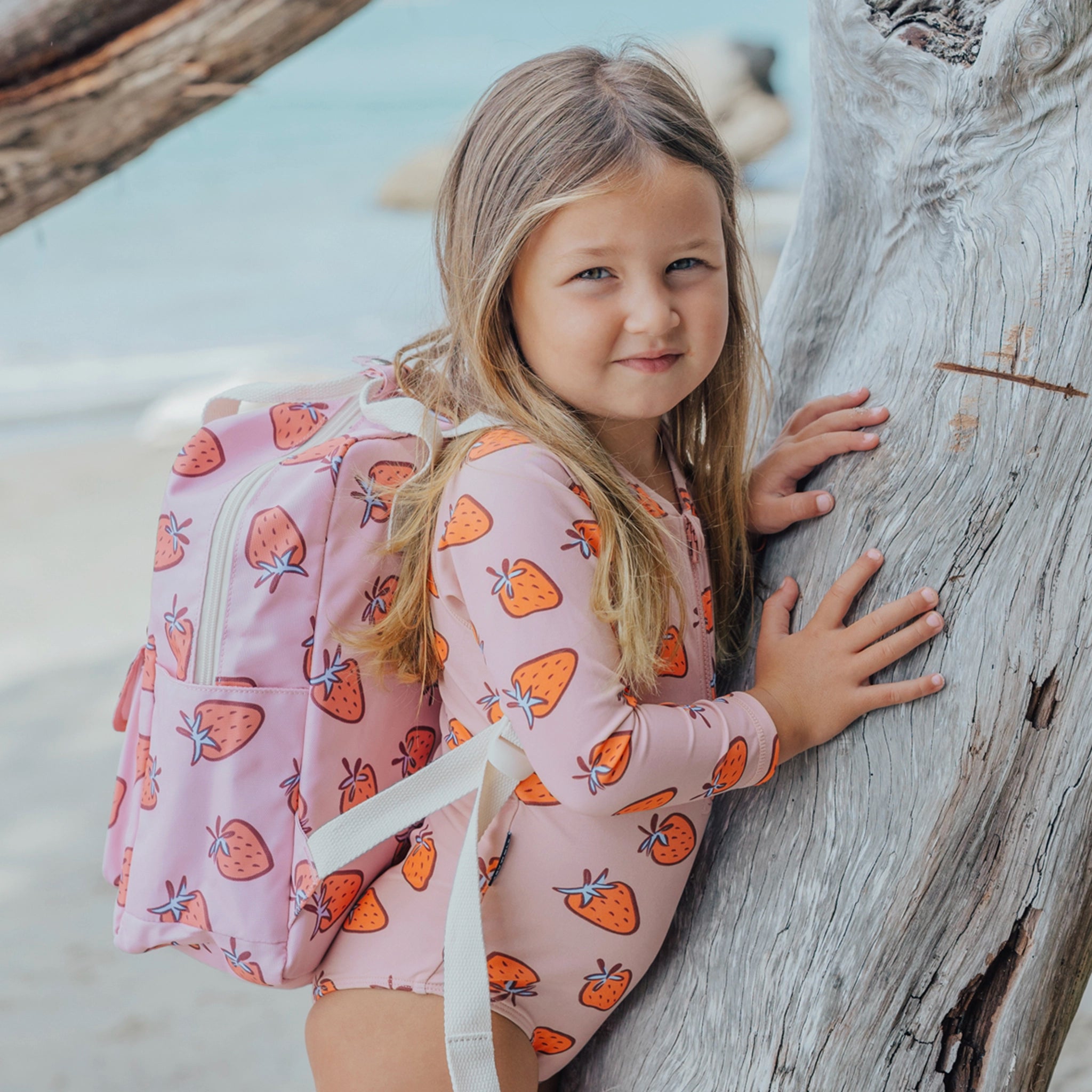 Young girl in a strawberry-patterned dress leaning against a wooden log on a beach.