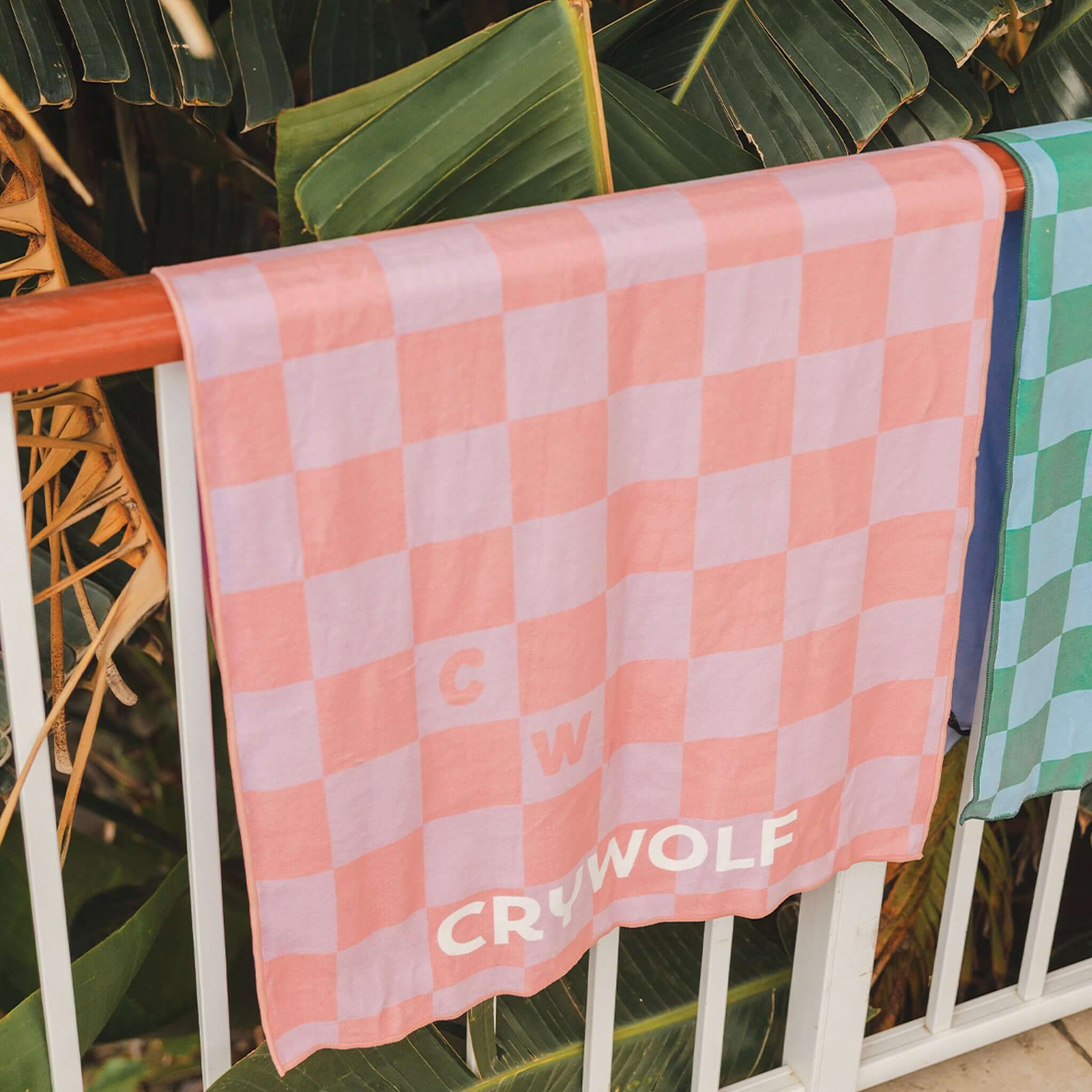 Crywolf Rapid Dry Towel - Flamingo Check hanging on a railing next to green towels, surrounded by tropical leaves.