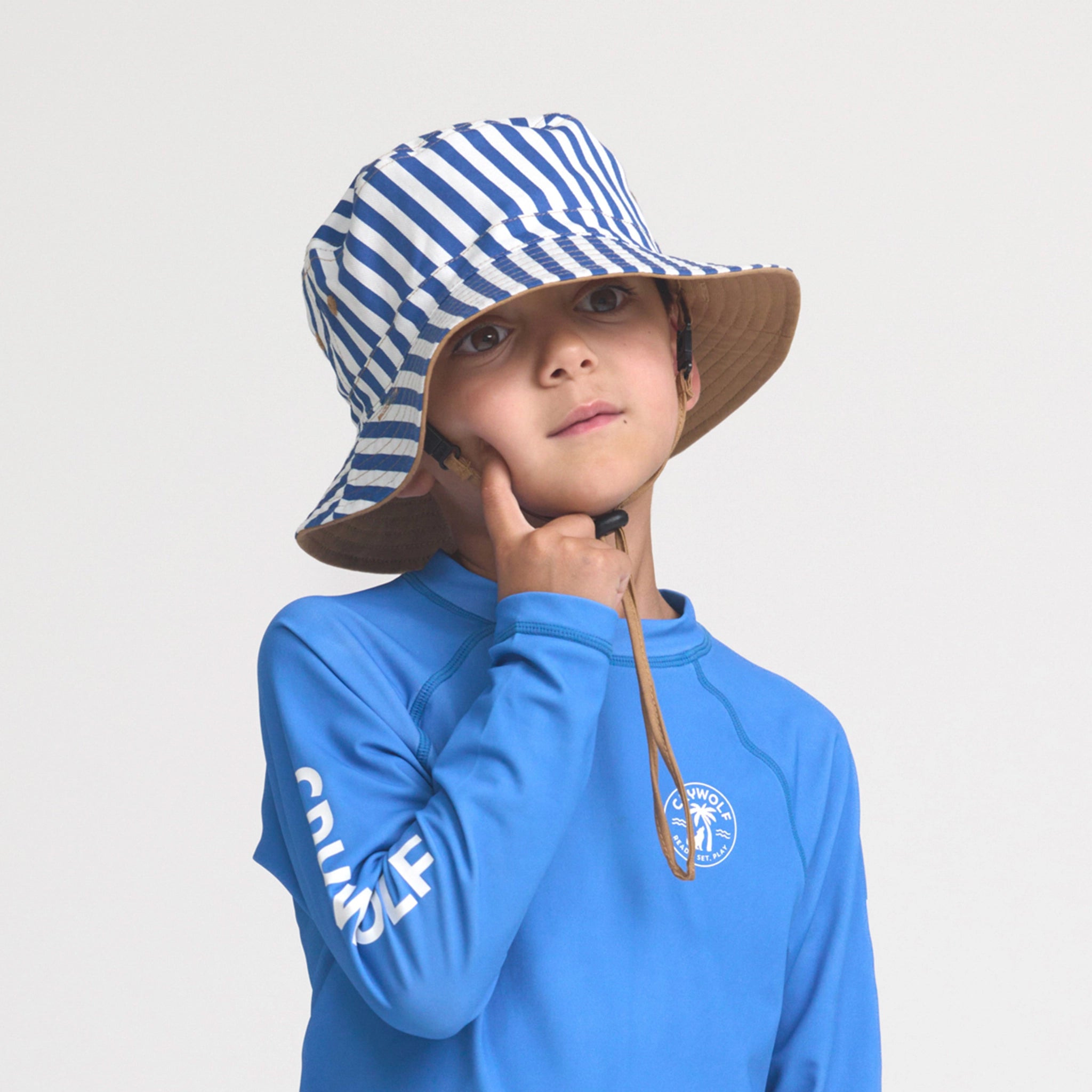 Child wearing a blue long-sleeve shirt with a logo and a striped bucket hat on a light gray background