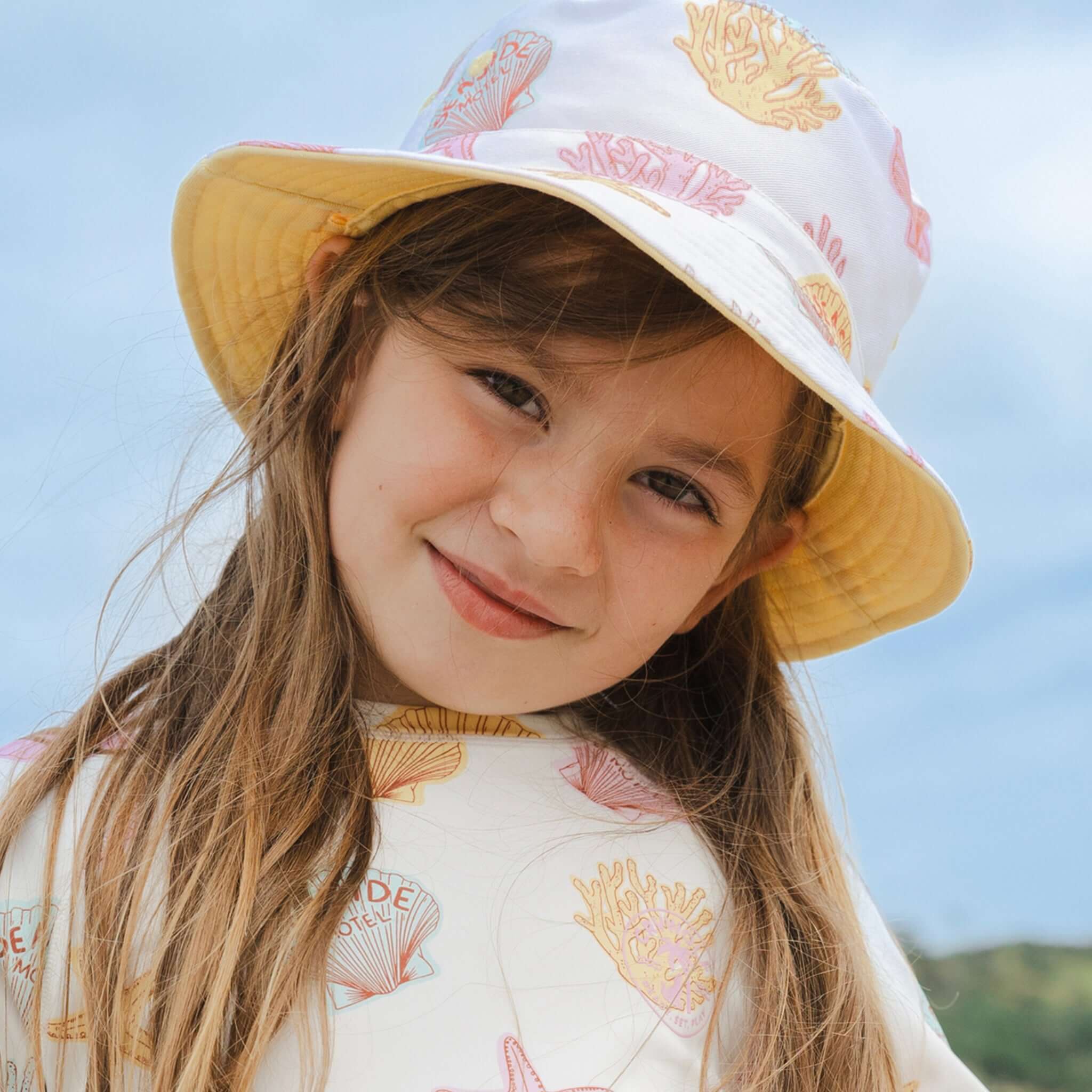 Girl wearing the Crywolf Reversible Bucket Hat - Shell, smiling outdoors with light blue background.