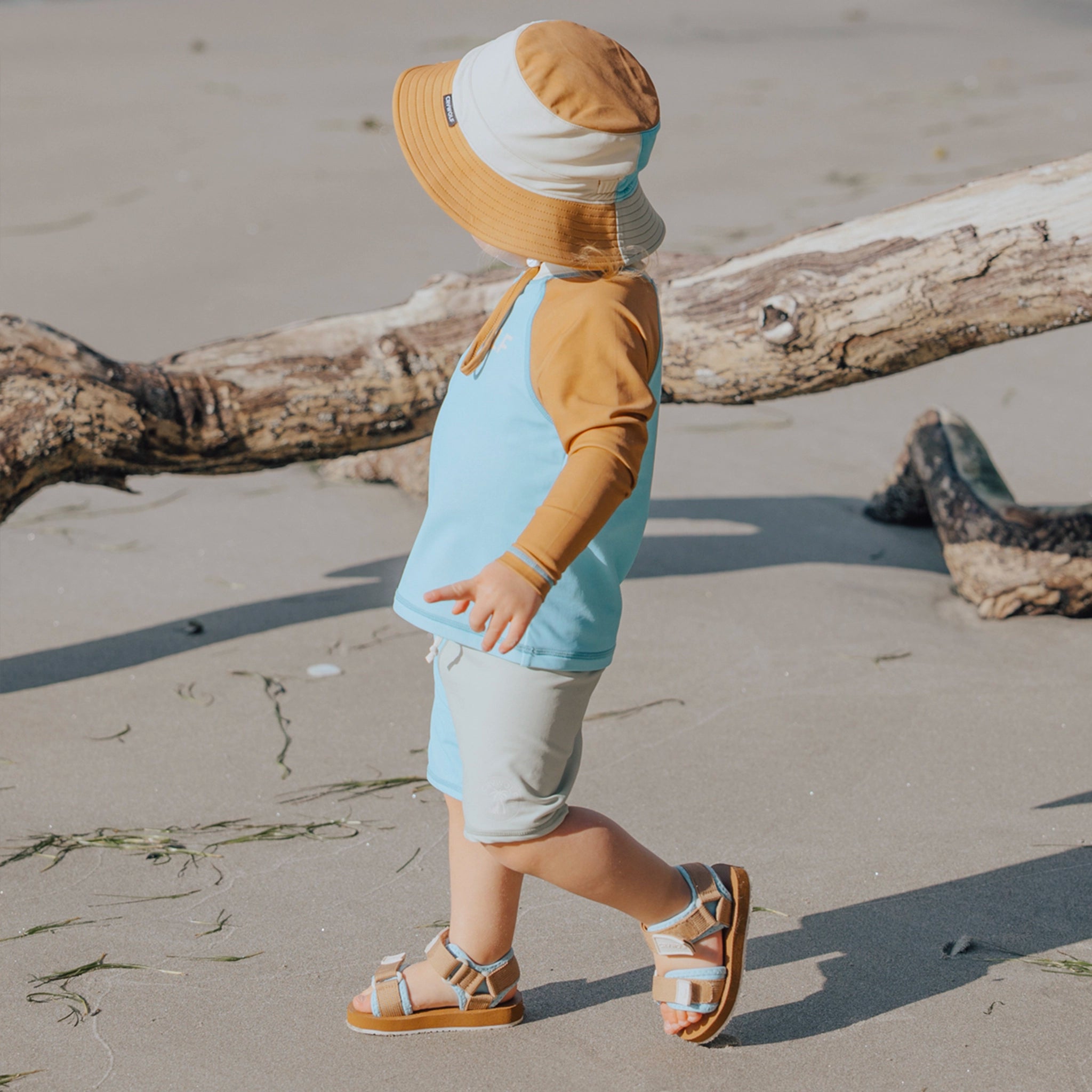 Child on a beach wearing a sun hat, blue shirt, and beige shorts.