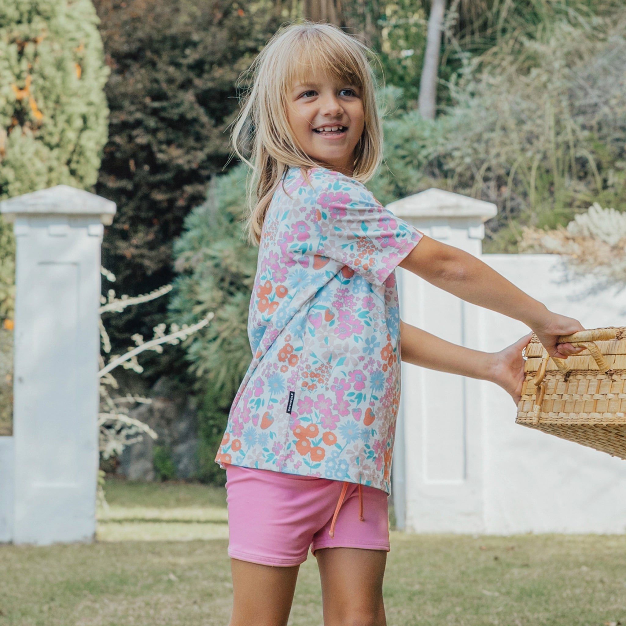 Young girl in a floral shirt and pink shorts holding a picnic basket outdoors.
