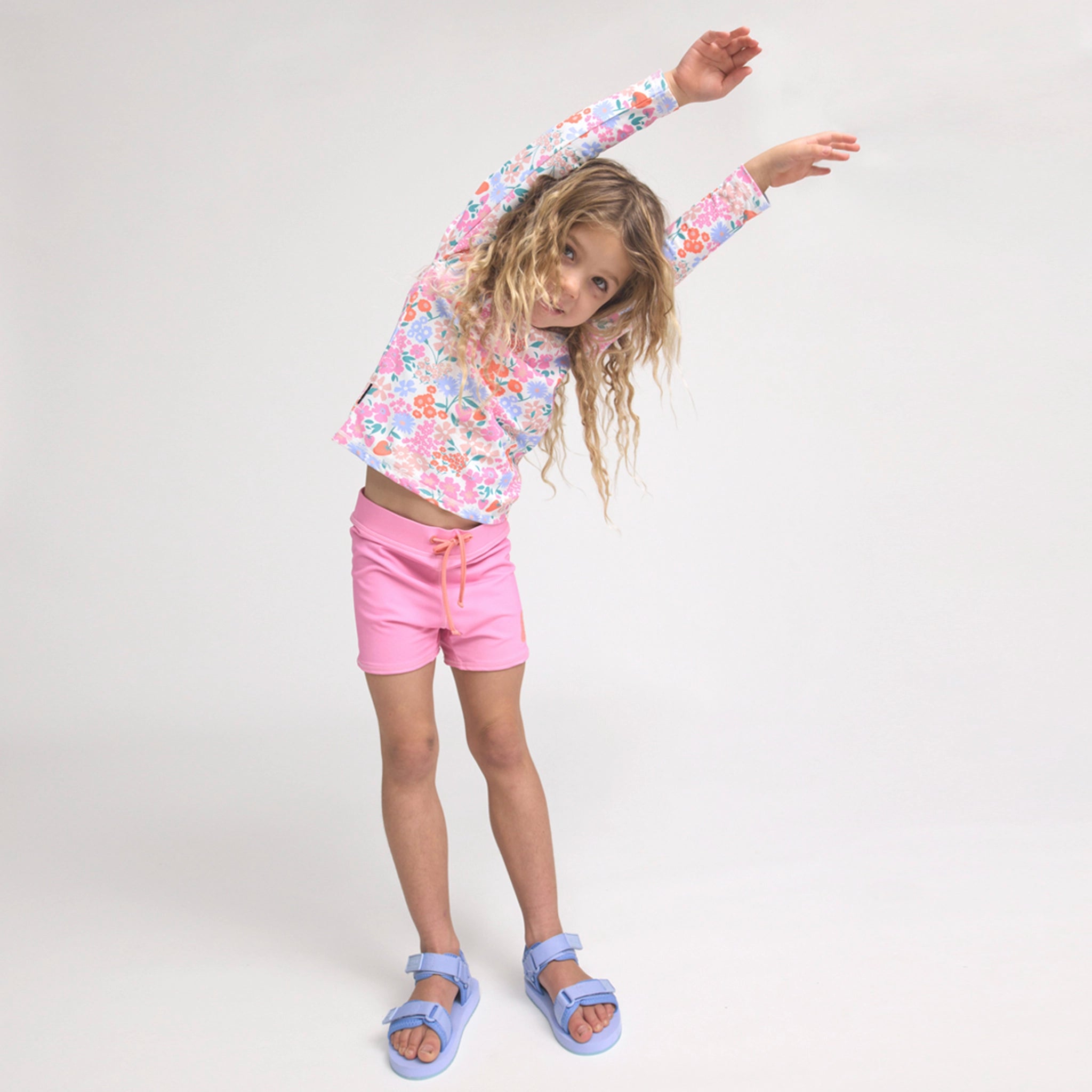 Young girl in a floral long-sleeve shirt and pink shorts standing against a white background