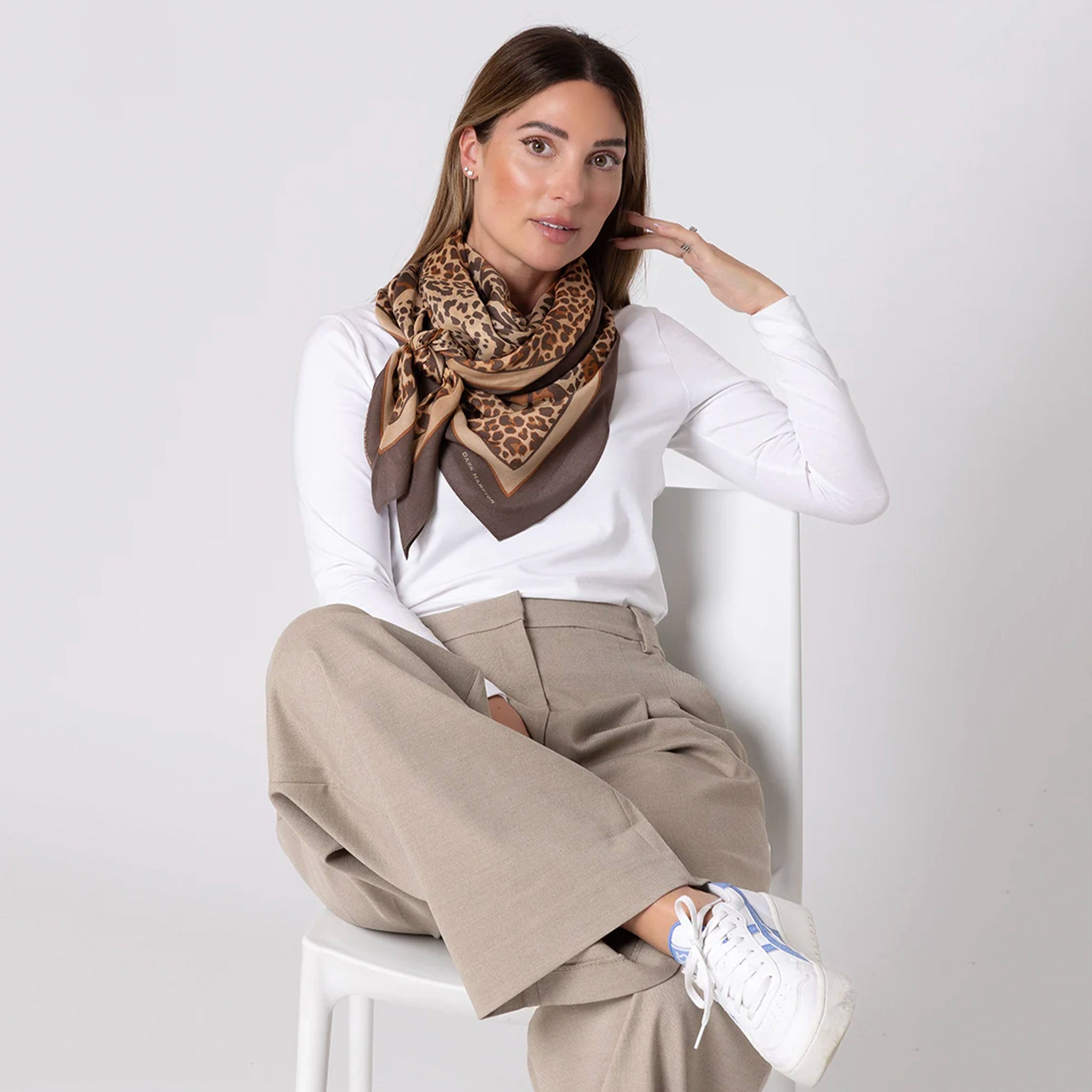 Woman wearing a leopard print scarf, white shirt, beige pants, and white sneakers on a light gray background
