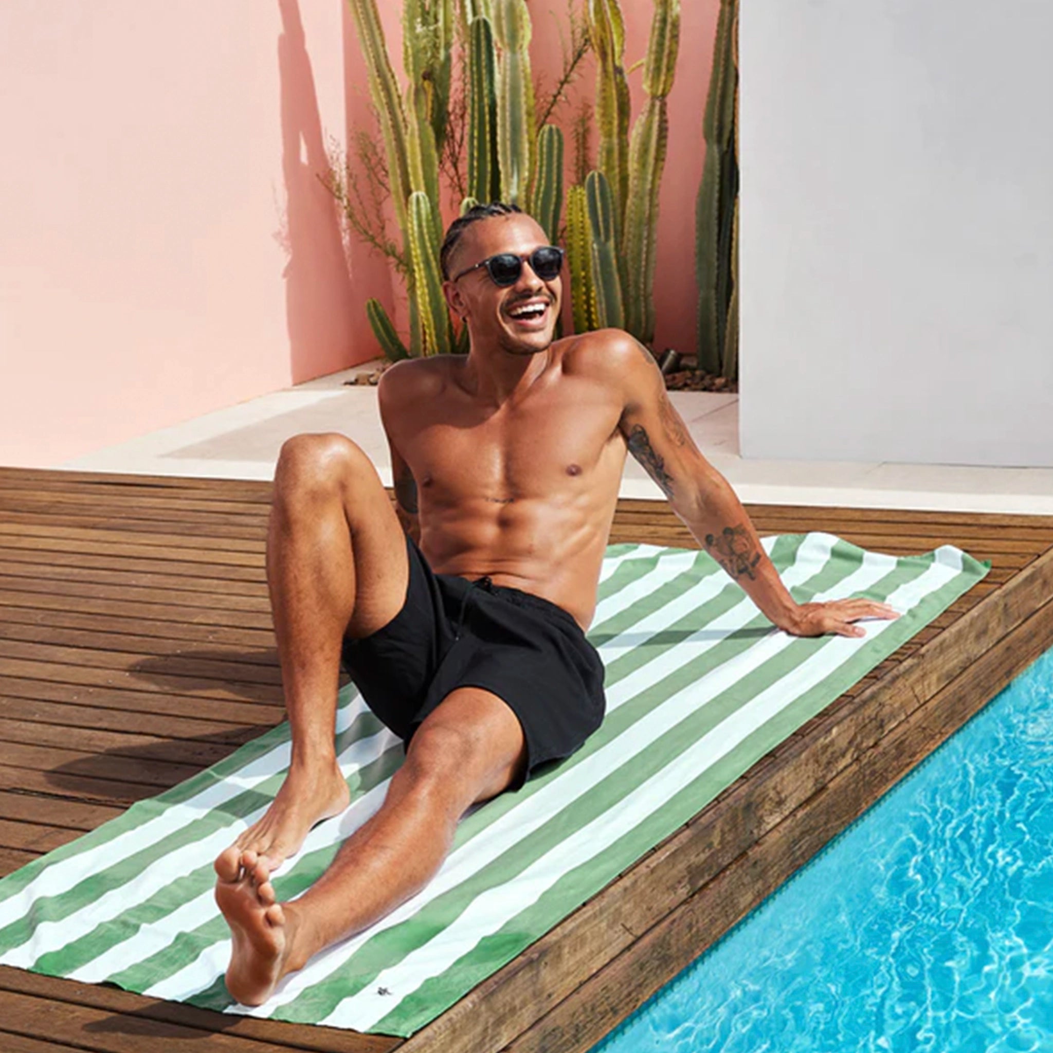 Man sitting on a green and white striped towel by a pool with cacti in the background