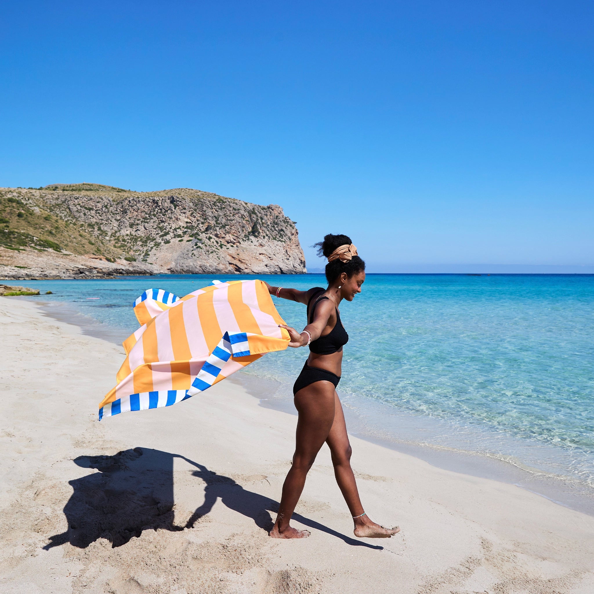 Woman holding a striped beach towel on a sandy beach with clear blue water and sky.