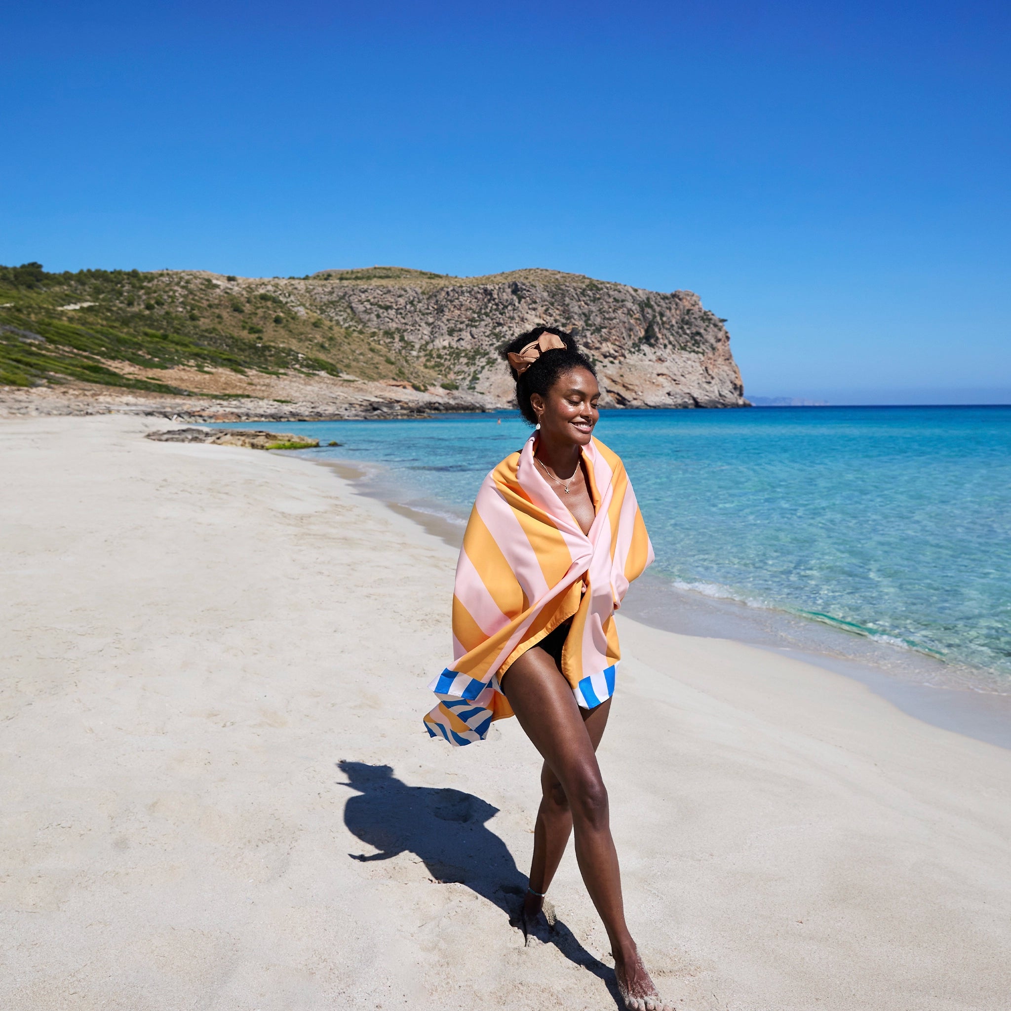 Woman wrapped in a colorful towel on a sandy beach with clear blue water and cliffs in the background