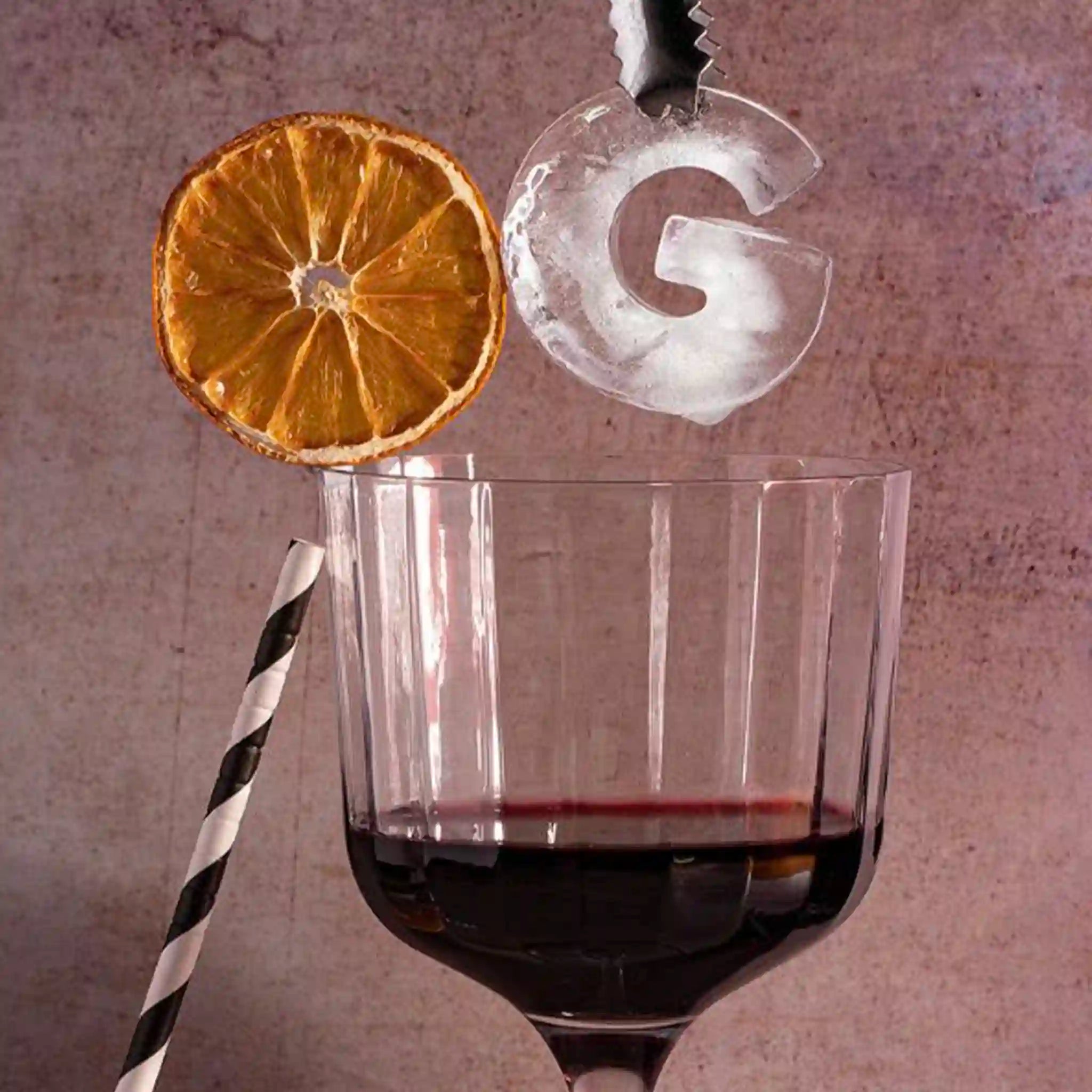 Glass of red wine with a dried orange slice and letter 'G' ice cube on a textured surface