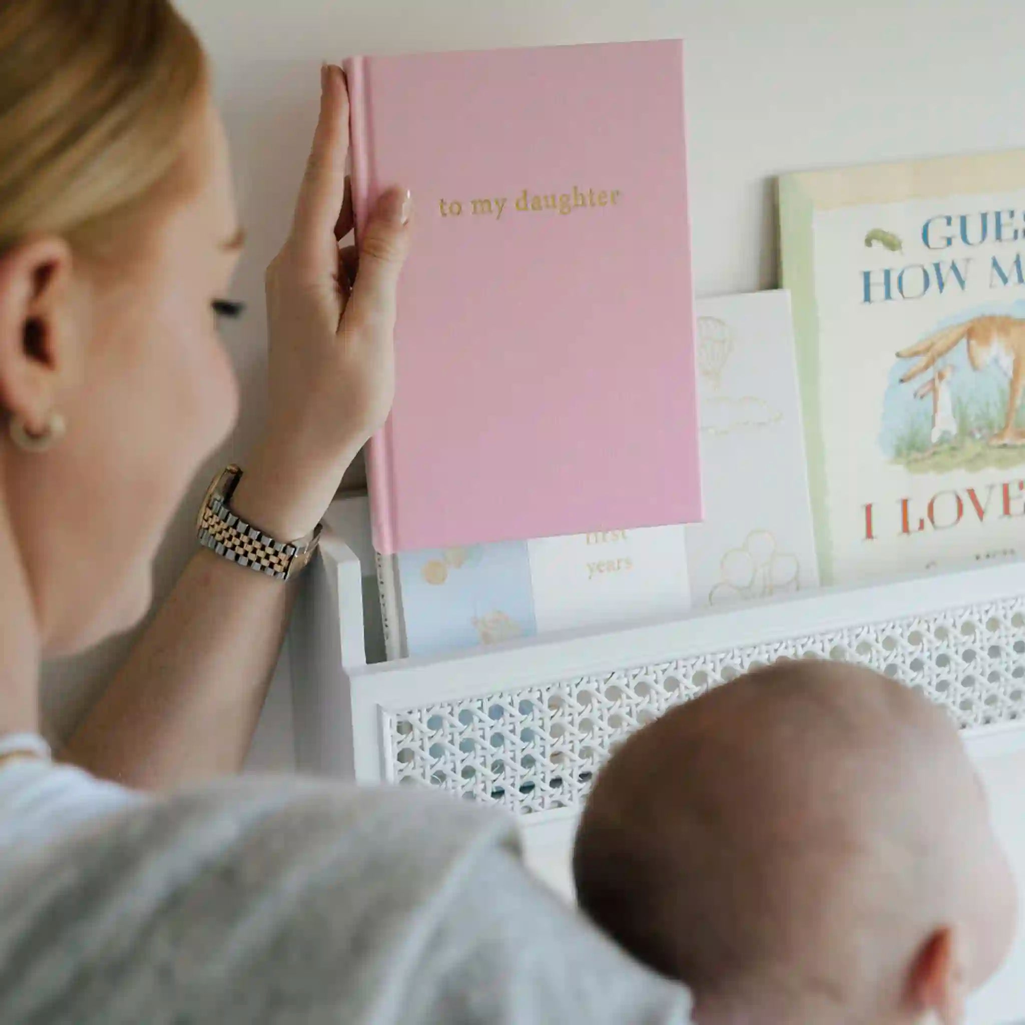 Woman reading a pink book titled 'to my daughter' to a baby in a nursery.