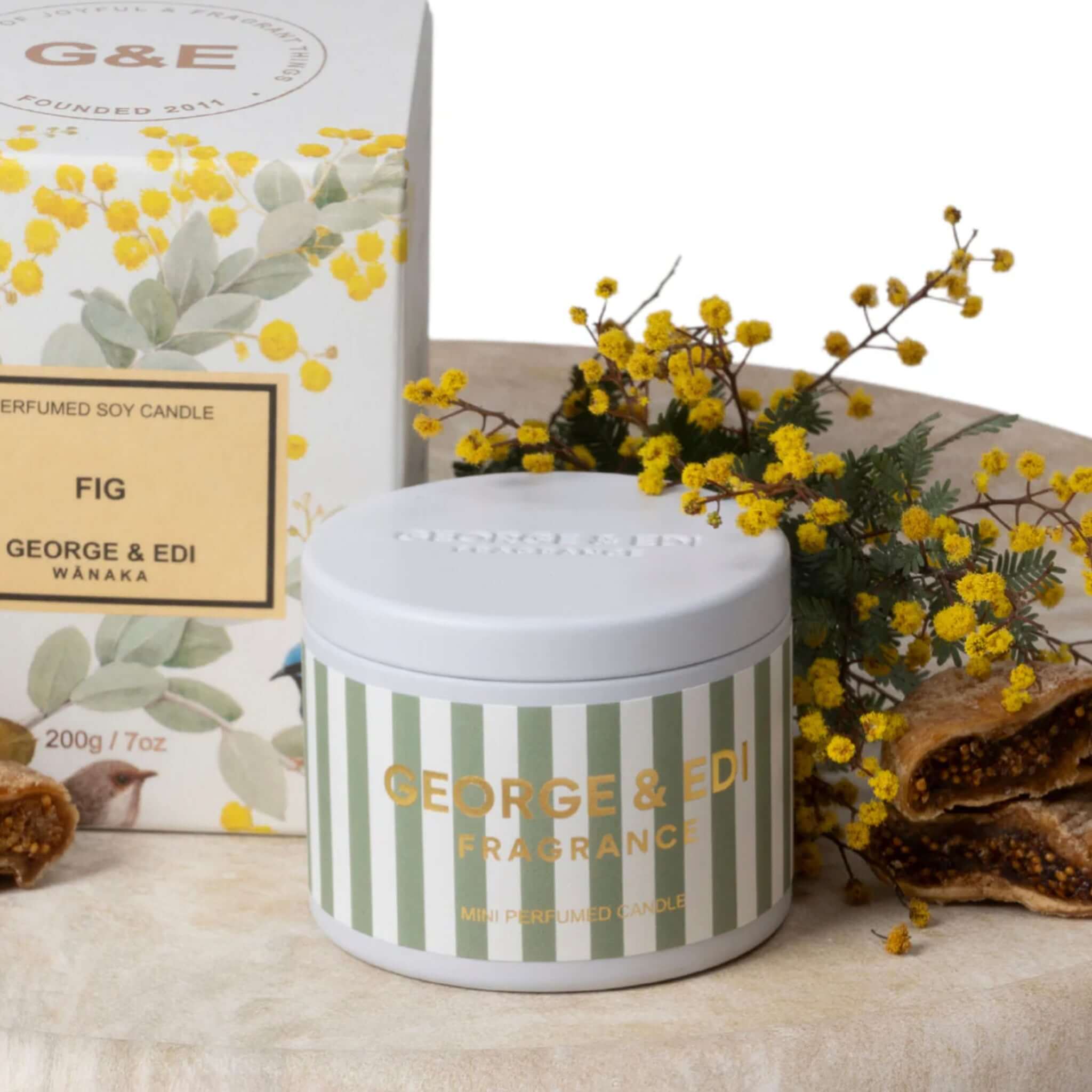 George & Edi Fragrance Mini Candle in a Tin - Fig on a wooden surface with flowers and herbs.