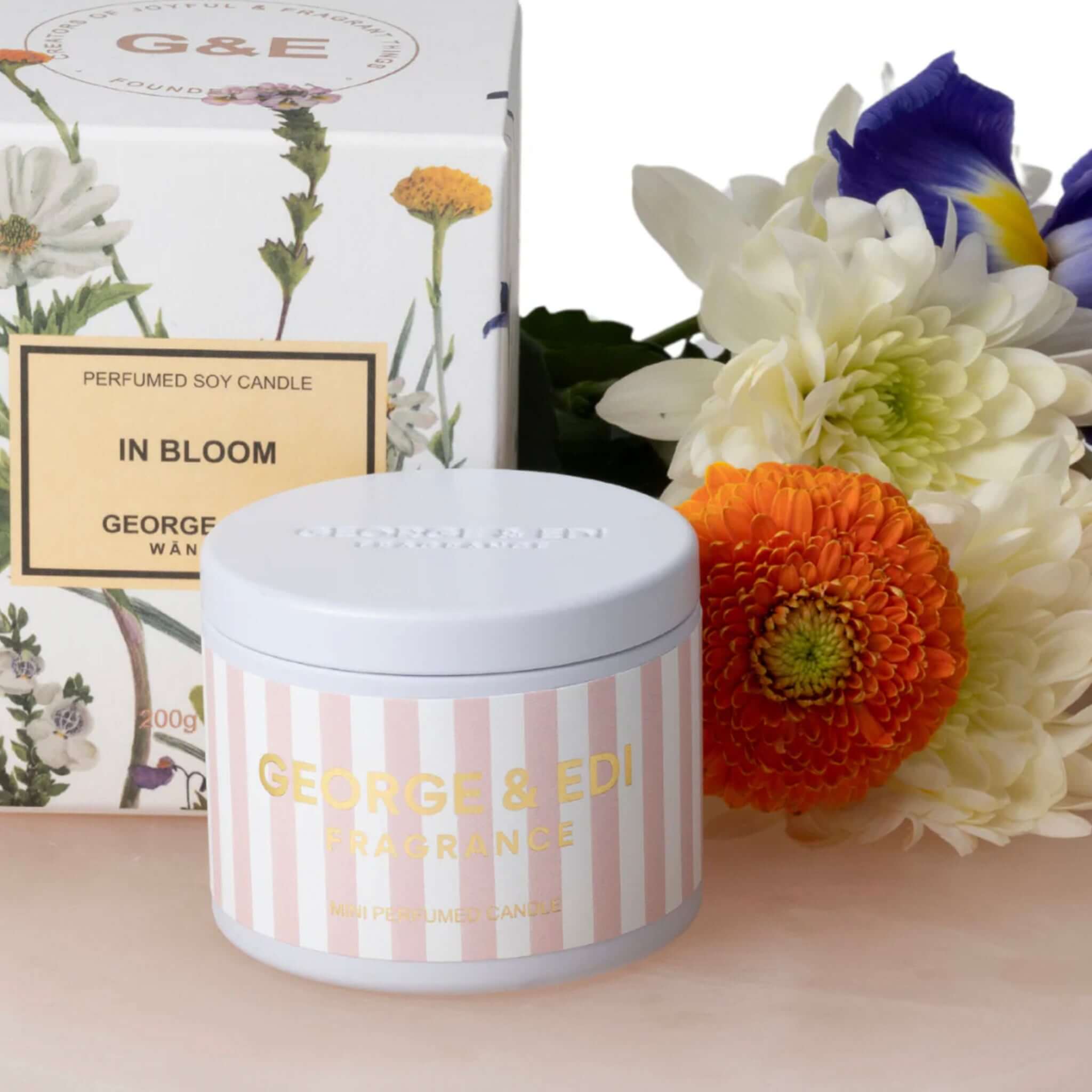 George & Edi Fragrance Mini Candle in a Tin - In Bloom against a floral backdrop, showcasing its elegant design.