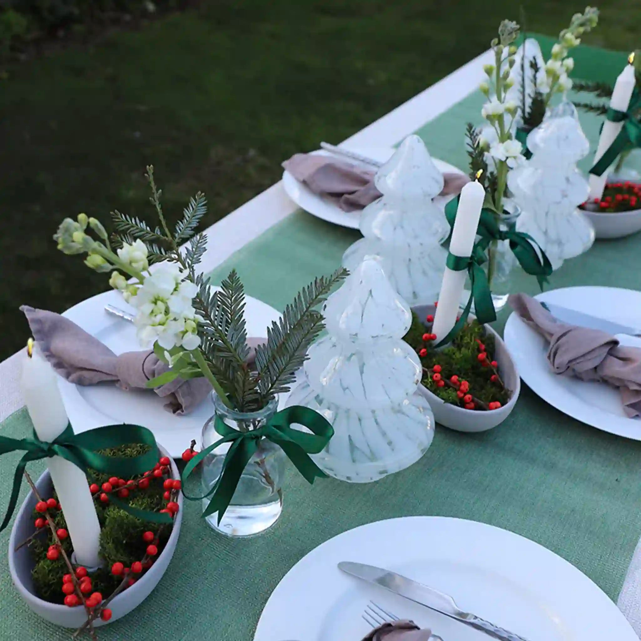 Decorative table setting with white candles, greenery, and red berries on a green tablecloth.