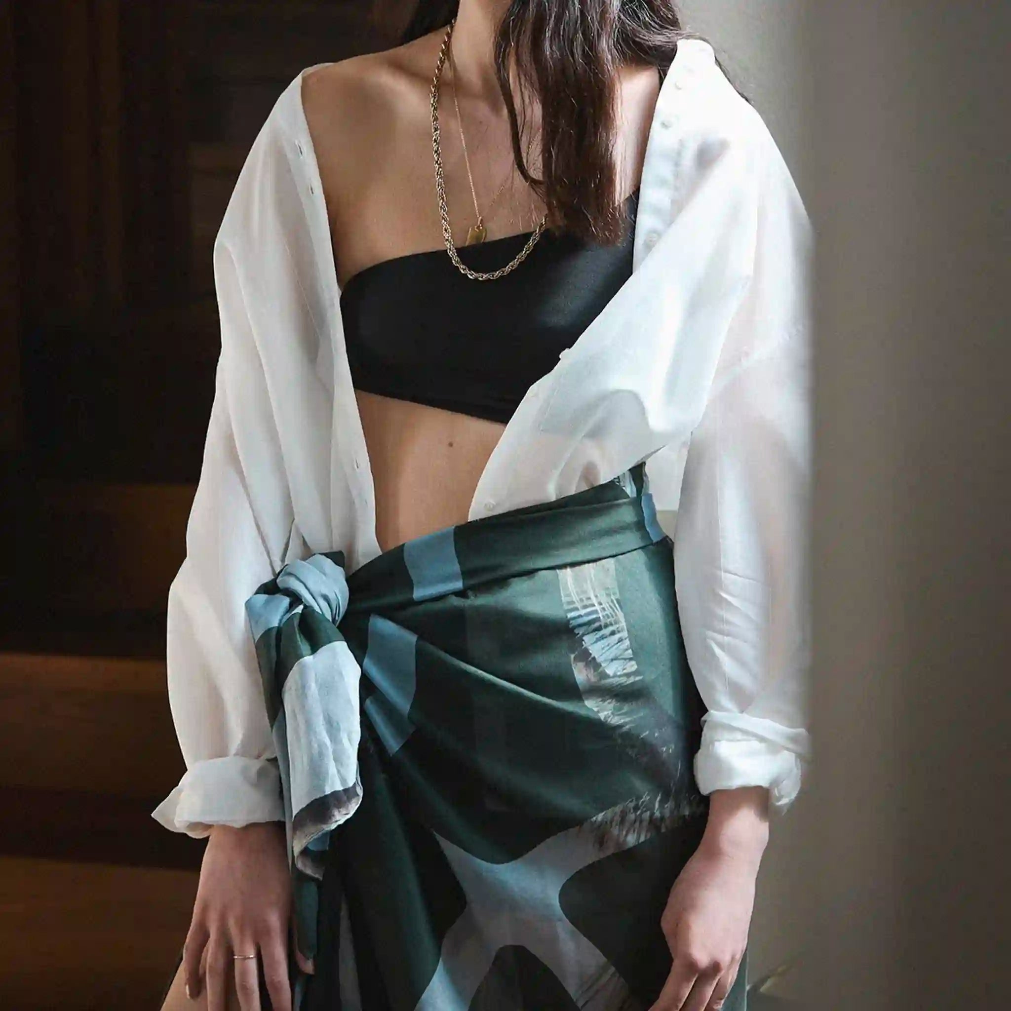 Person wearing a white shirt over a black bralette with a green and gray patterned skirt.