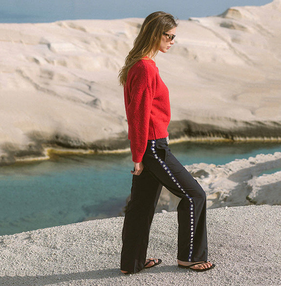 A woman stands outdoors by a turquoise body of water with pale rock formations in the background, wearing a bright red knit sweater, black wide-leg pants with a decorative white and blue side stripe, and dark sandals.