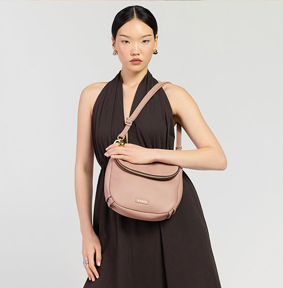 A model wearing a dark brown halter dress holds a dusty pink crescent-shaped shoulder bag with gold hardware against a light gray studio background.