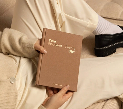 Person holding a brown notebook with 'Two thousand Twenty Six' text, sitting on a beige couch.