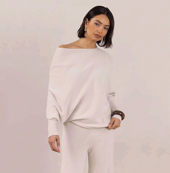 A woman modeling a light cream off-the-shoulder knit sweater and matching knit pants against a soft beige studio background.