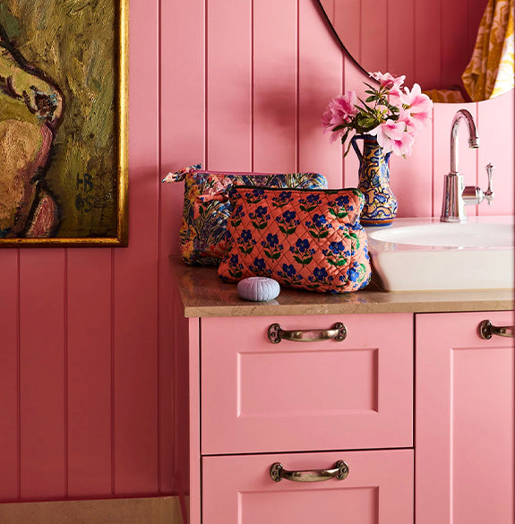 Pink bathroom with floral bags on a counter, flowers in a vase, and a sink.