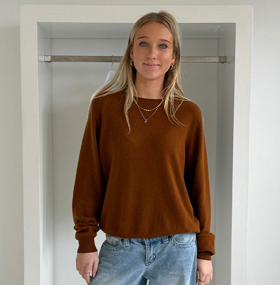 Woman wearing a brown sweater and blue jeans standing in a neutral-colored room.