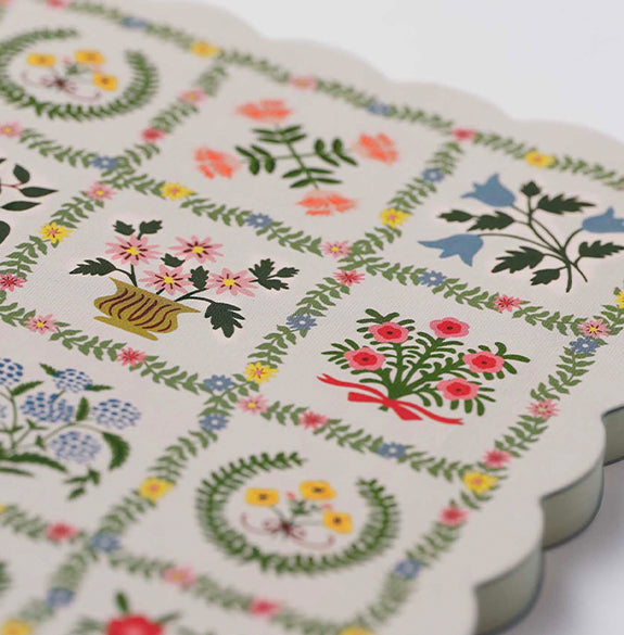 A close-up of a scalloped fabric item, likely a napkin or coaster, featuring a vintage-style patchwork design with floral and botanical motifs in green, pink, yellow, and blue on an off-white background.