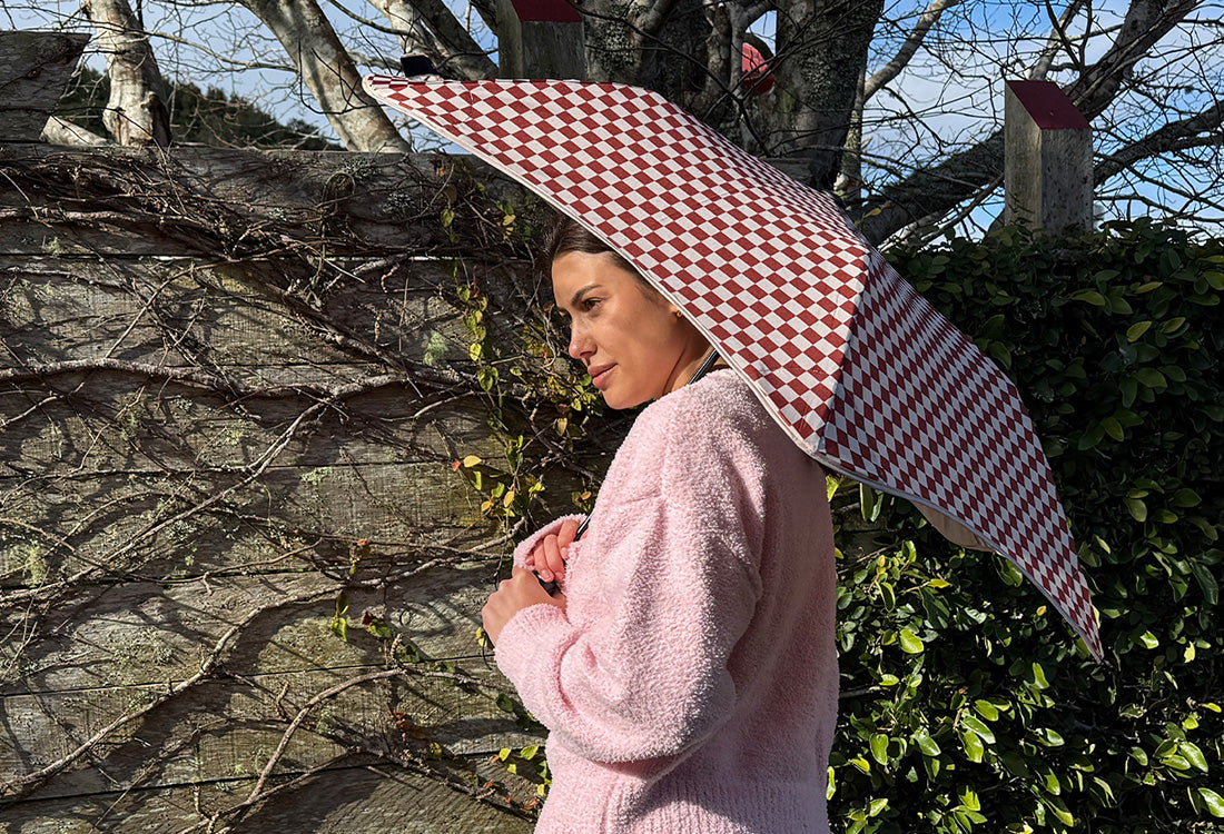 A woman in a fuzzy light pink sweater stands outdoors under a large open umbrella with a red and white checkered pattern, set against a background of ivy-covered trees.