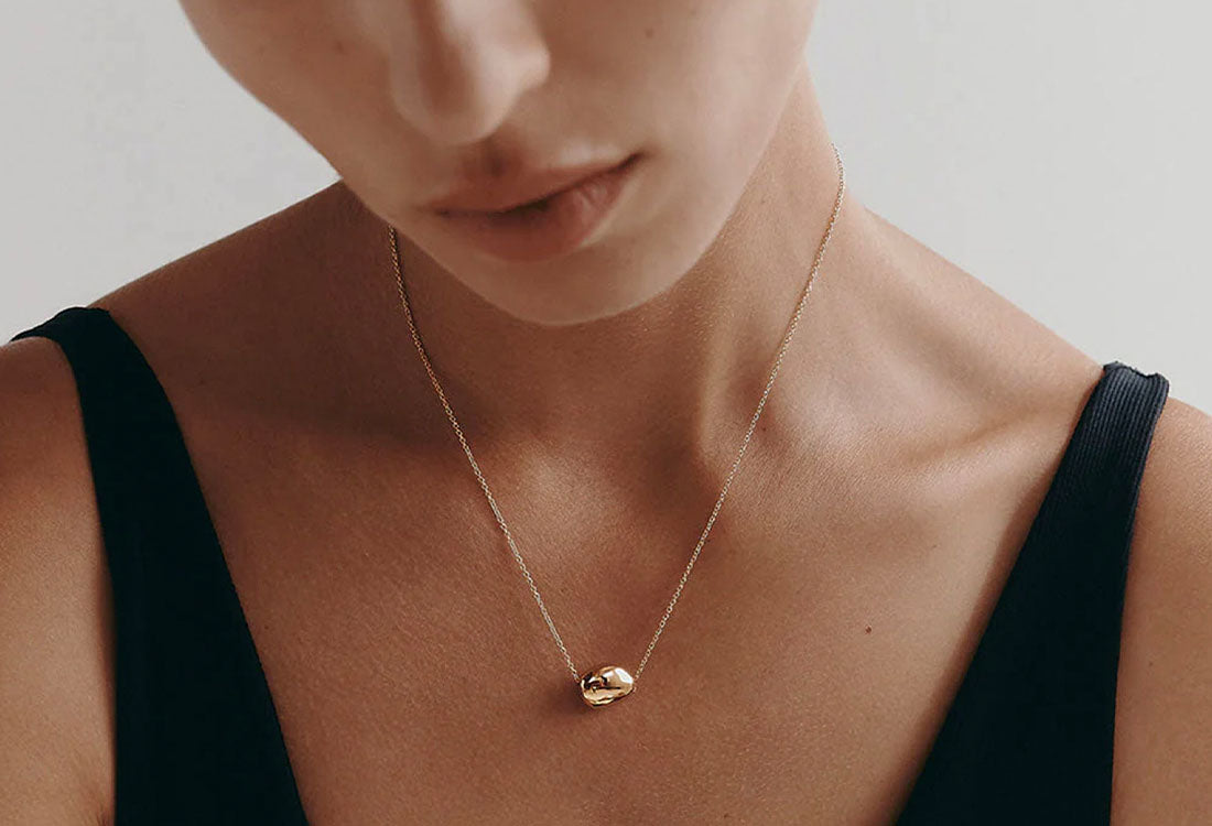 Close-up of a person wearing a gold necklace with a small pendant against a neutral background