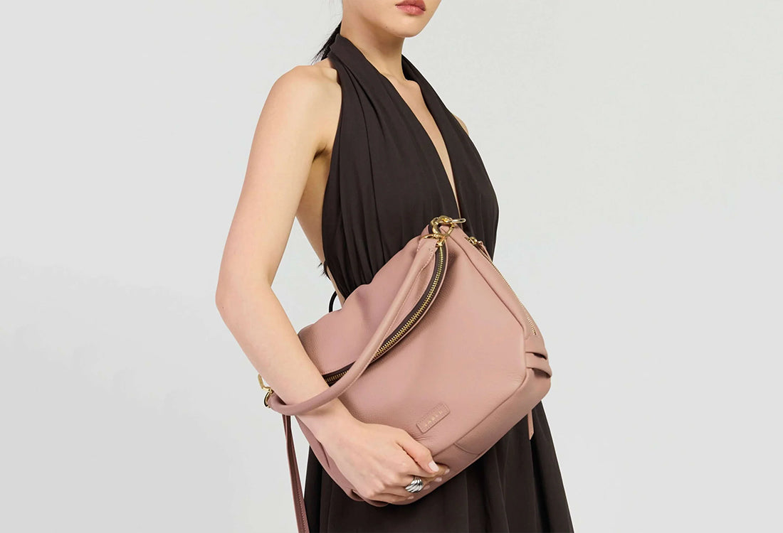 A woman wearing a black halter dress carries a dusty pink leather shoulder bag with gold-tone hardware against a light gray studio background.