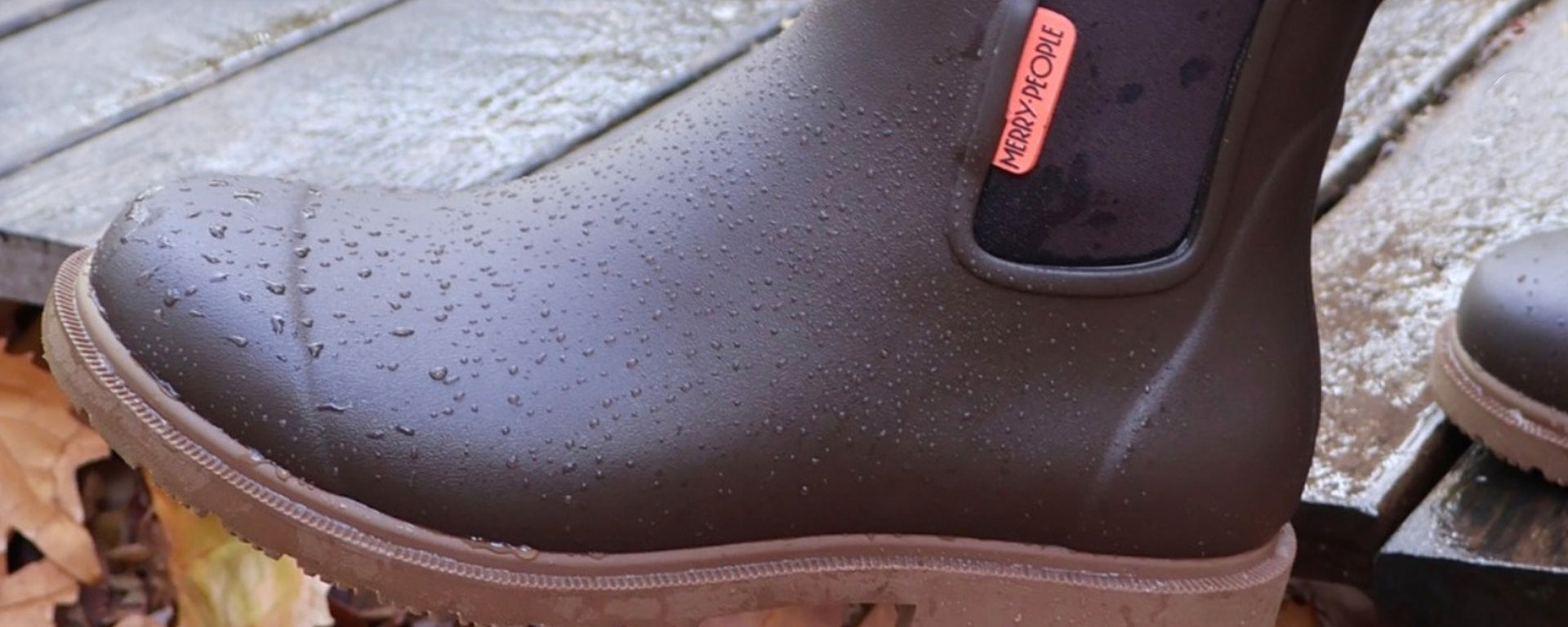 A close-up of a dark brown rubber rain boot covered in water droplets, resting on wet wooden decking outdoors. A small pink tag on the side reads 'HARPER-FOX'.