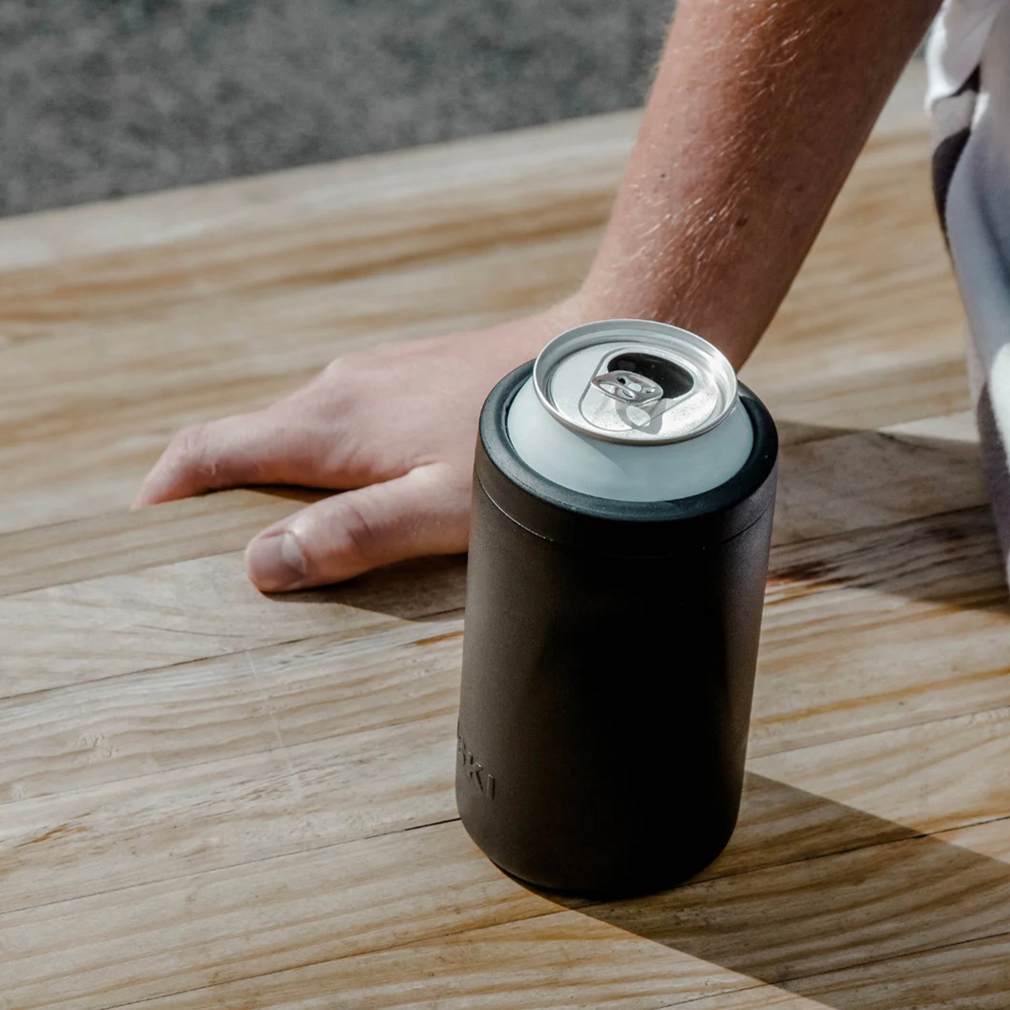 Person holding a can with a black can cooler on a wooden surface