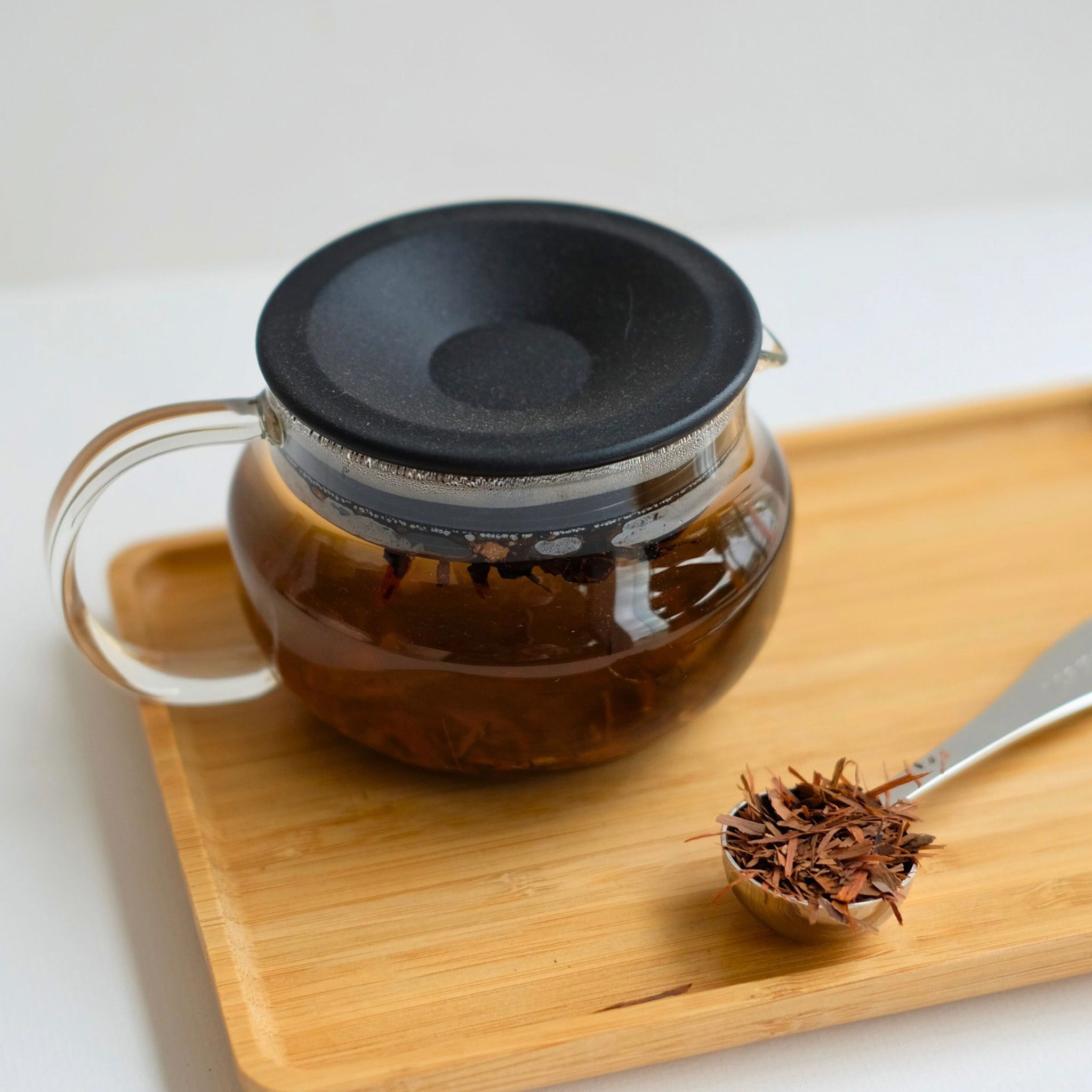 Glass teapot with black lid on a wooden tray, accompanied by a spoonful of tea leaves.