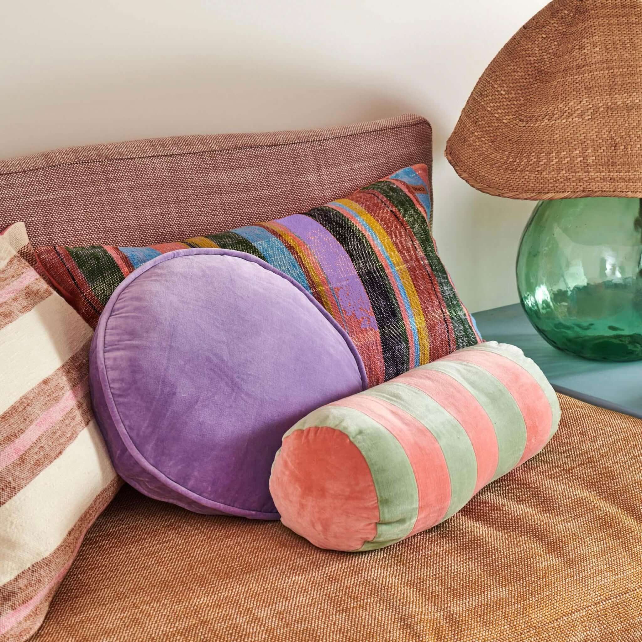 Alt Tag: Kip & Co Velvet Pea Cushion - Lilac round lilac velvet cushion styled on sofa with striped bolster and patterned pillow